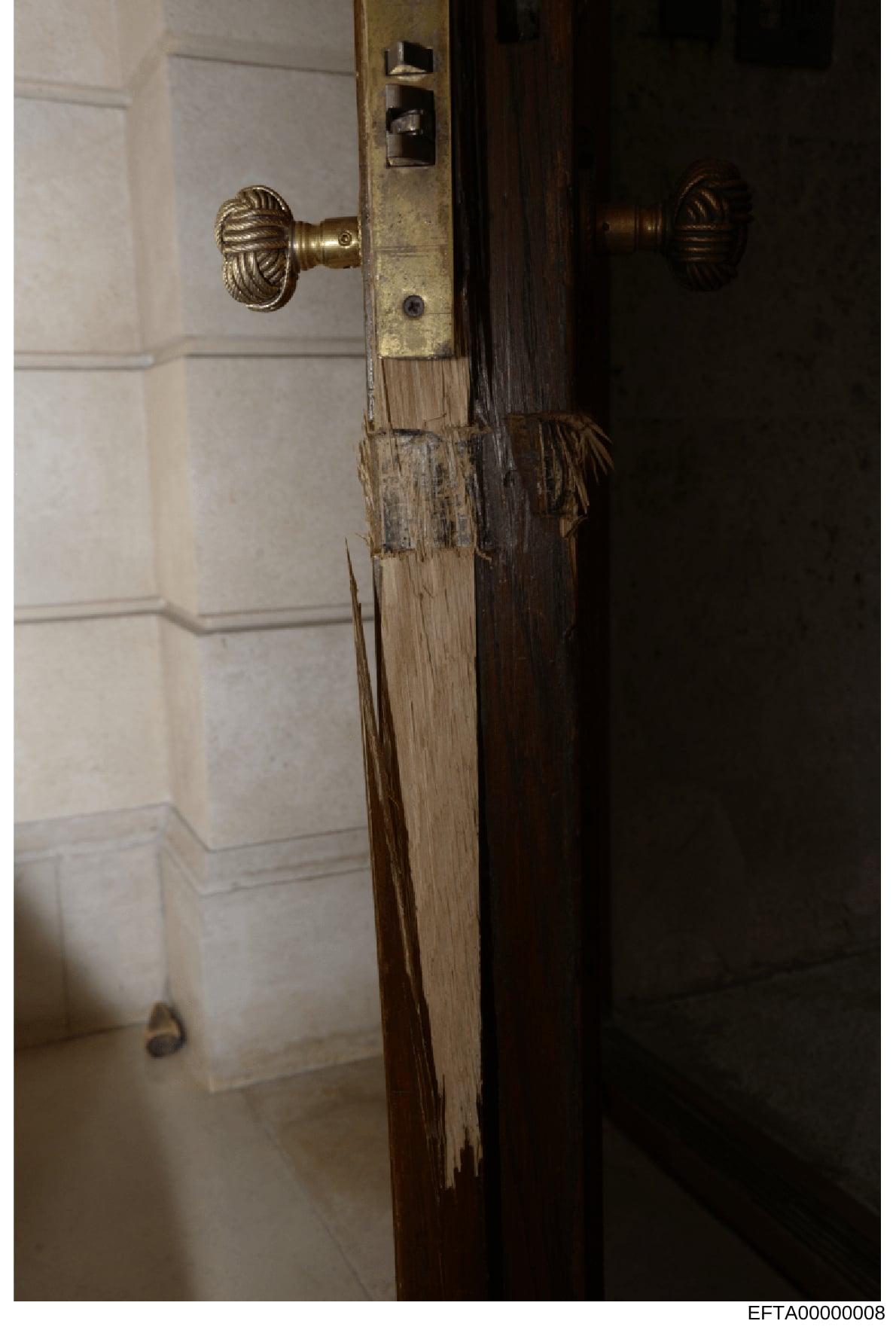 This is an evidence photograph showing the interior detail of a wooden door frame and brass door hardware, likely from one of Epstein's properties. The image captures the door's wooden frame with visible wear and deterioration, brass ornamental door