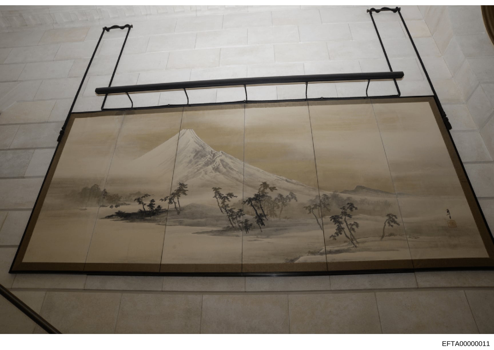 This photograph documents a framed piece of Asian artwork, likely a painting or print on silk or paper depicting a misty mountain landscape with a snow-capped peak, trees, and water. The artwork is mounted in a traditional frame with horizontal suppo