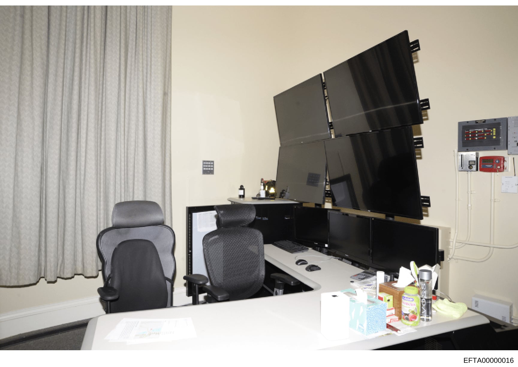 This is a photograph of a modern office space featuring multiple computer monitors mounted on a wall, ergonomic office chairs, and what appears to be a security or monitoring station. The setup includes surveillance or control room-style equipment wi
