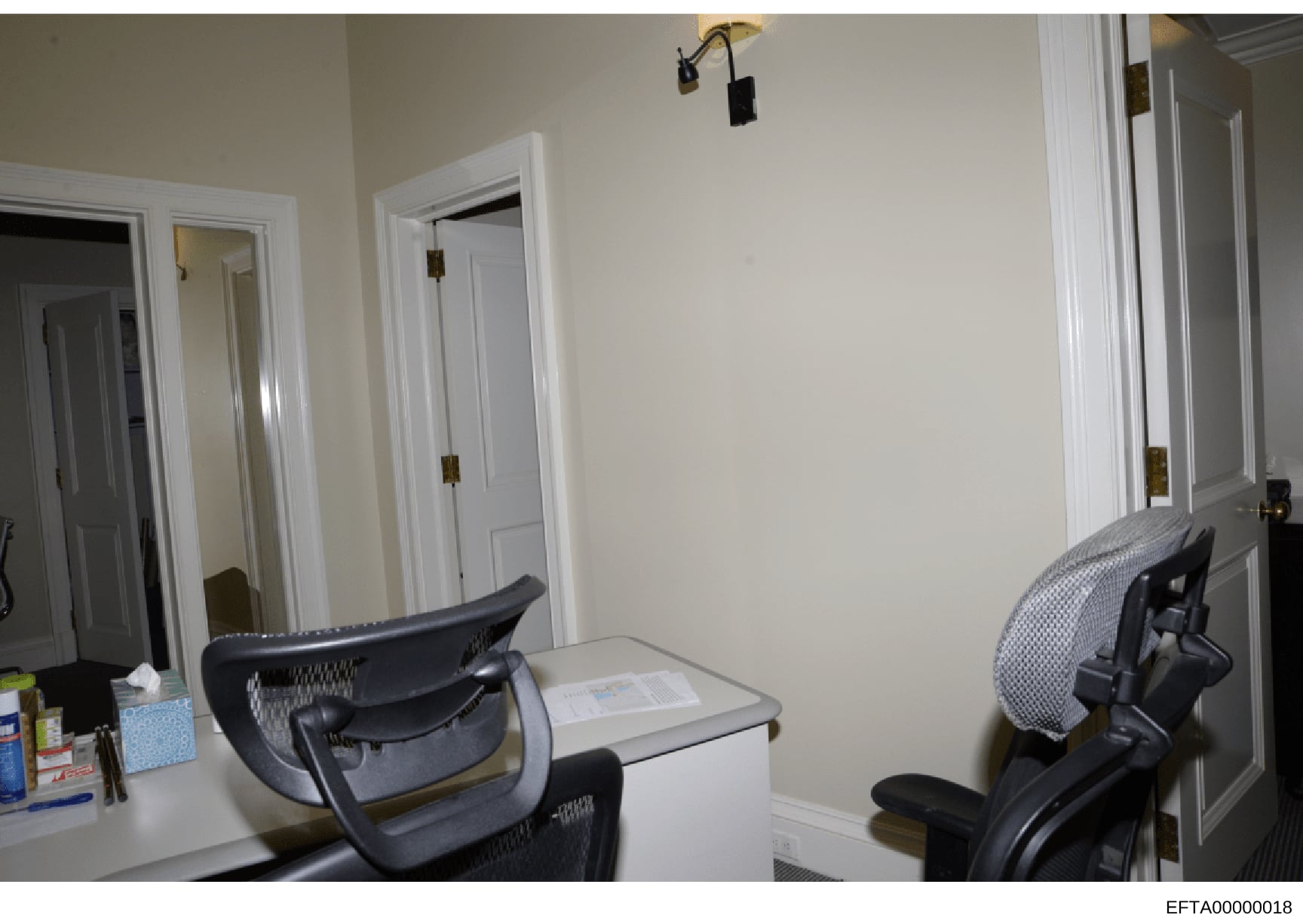 This is a photograph of an interior room, likely an office or bedroom space, showing multiple white doors with brass hardware, beige walls, a desk with office chairs, and basic furnishings. The image appears to be documentation of a property interior