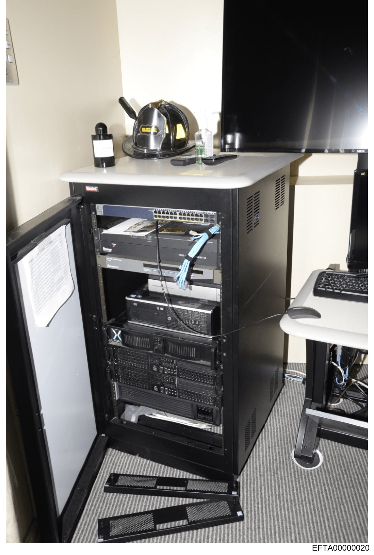 This is a photograph of a black server rack cabinet containing networking and computing equipment, located in an office space. The cabinet holds multiple rack-mounted devices including a network switch and various servers. A firefighter helmet sits a