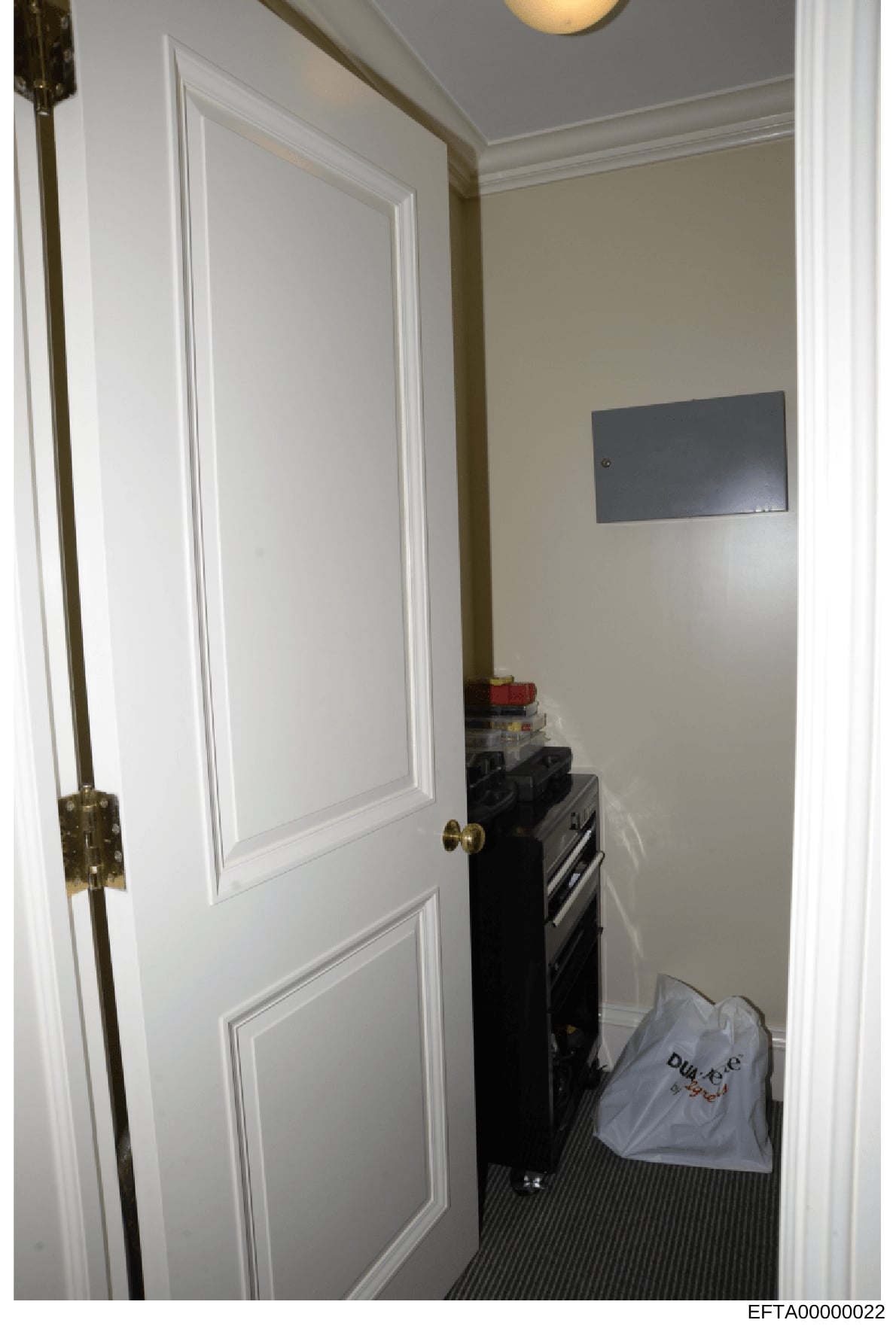 This is an evidence photograph showing an interior room space, featuring a white two-panel door with brass hinges, a mounted flat-screen monitor on the wall, and various items including what appears to be a black device or equipment and a light-color