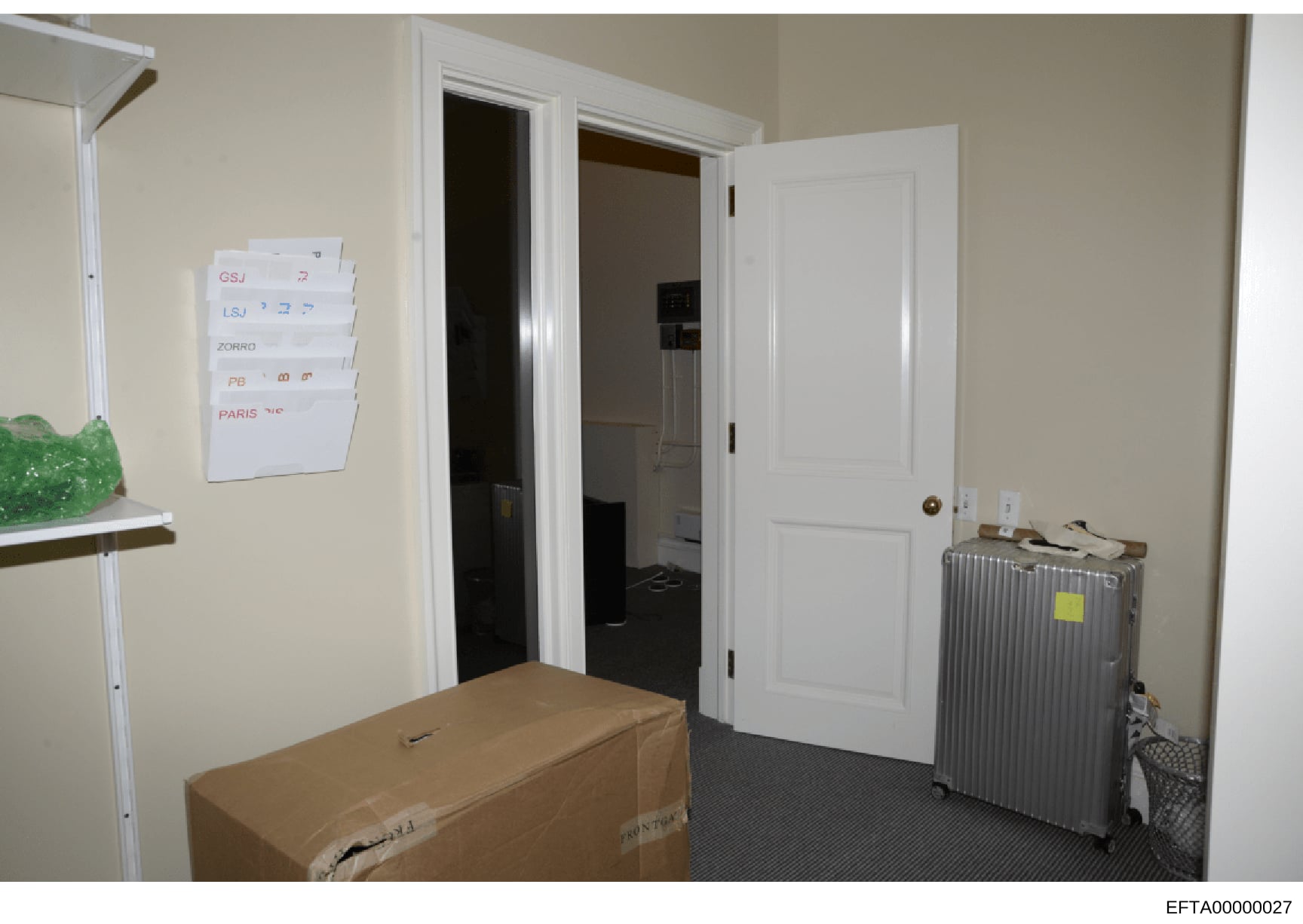 This photograph shows an interior room with organized travel documentation posted on the wall, including labels for various locations (GSJ, LSJ, Zorro, PB, Paris). A cardboard box and metal suitcase with a yellow tag are visible on the floor. The ima