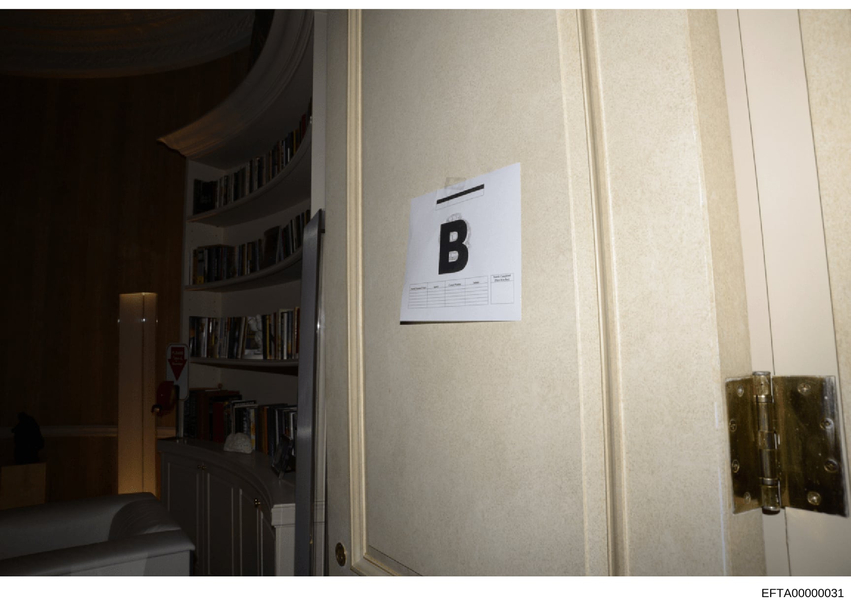 Evidence photograph documenting an interior space with built-in bookshelves and a white evidence marker labeled 'B' affixed to the wall. This appears to be a crime scene or property documentation photo, likely taken during the investigation of one of
