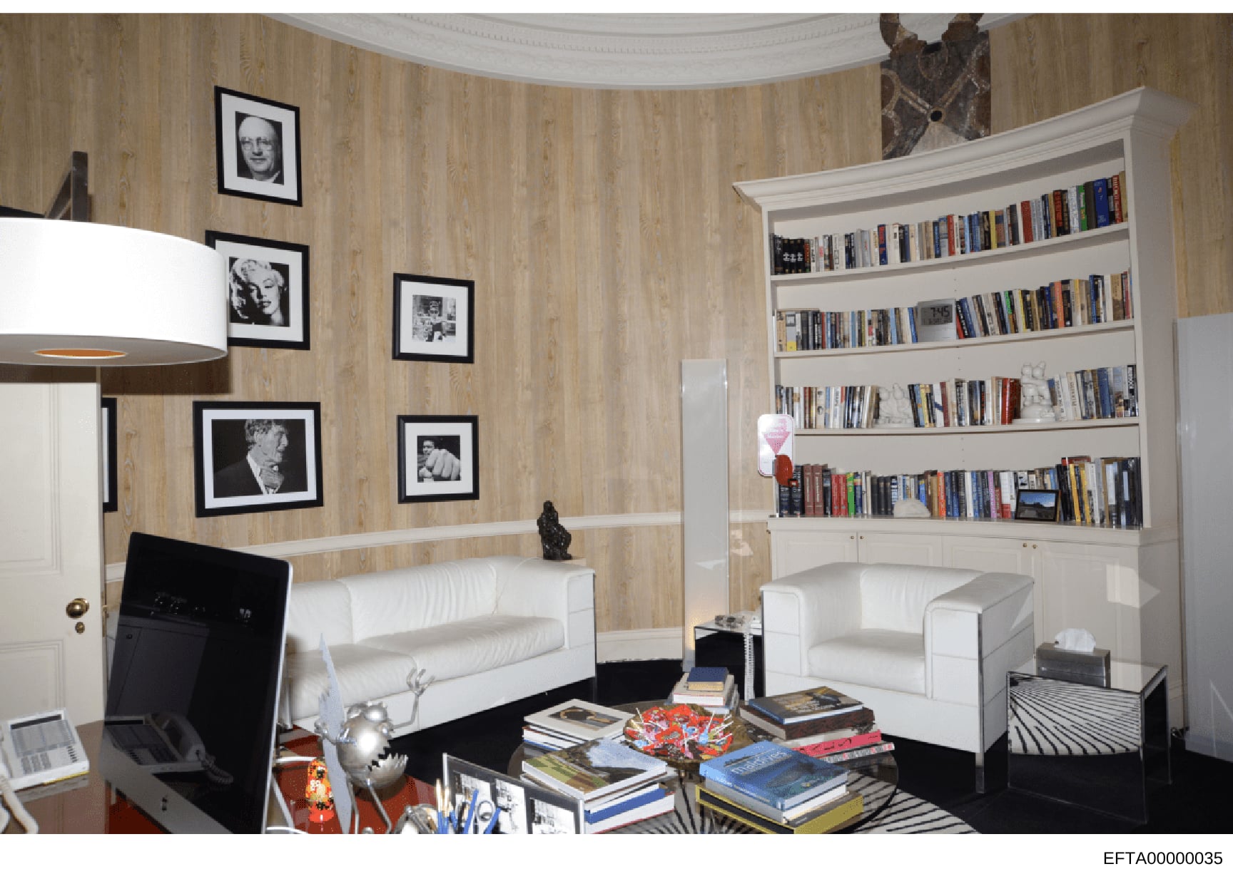 This is a photograph of an elegantly furnished residential interior space, featuring contemporary artwork, extensive bookshelves, and high-end furnishings. The room displays framed black-and-white portrait photographs on wood-textured walls, a curved