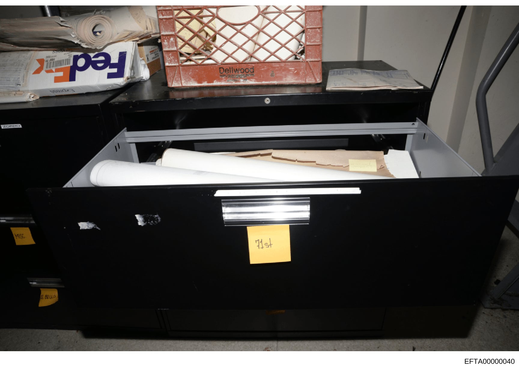 This photograph shows a black evidence storage container or filing drawer that is open, displaying stored documents, rolled blueprints or plans, and various materials. Yellow evidence tags are visible on the exterior, suggesting this is an official e