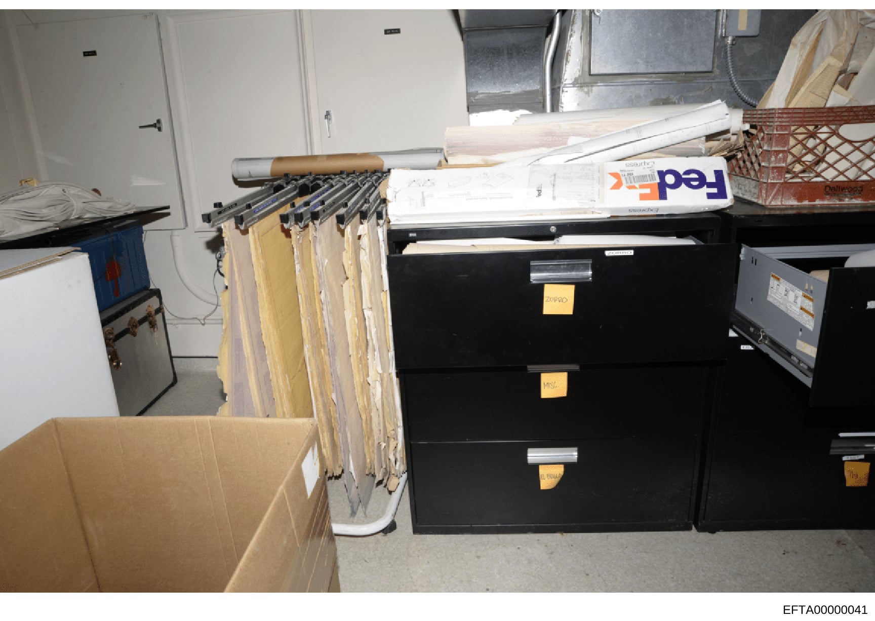 This photograph shows a storage room or evidence facility containing multiple black filing cabinets with yellow identification tags, hanging file folders on a rod system, cardboard boxes, and stacked documents including what appears to be FedEx packa