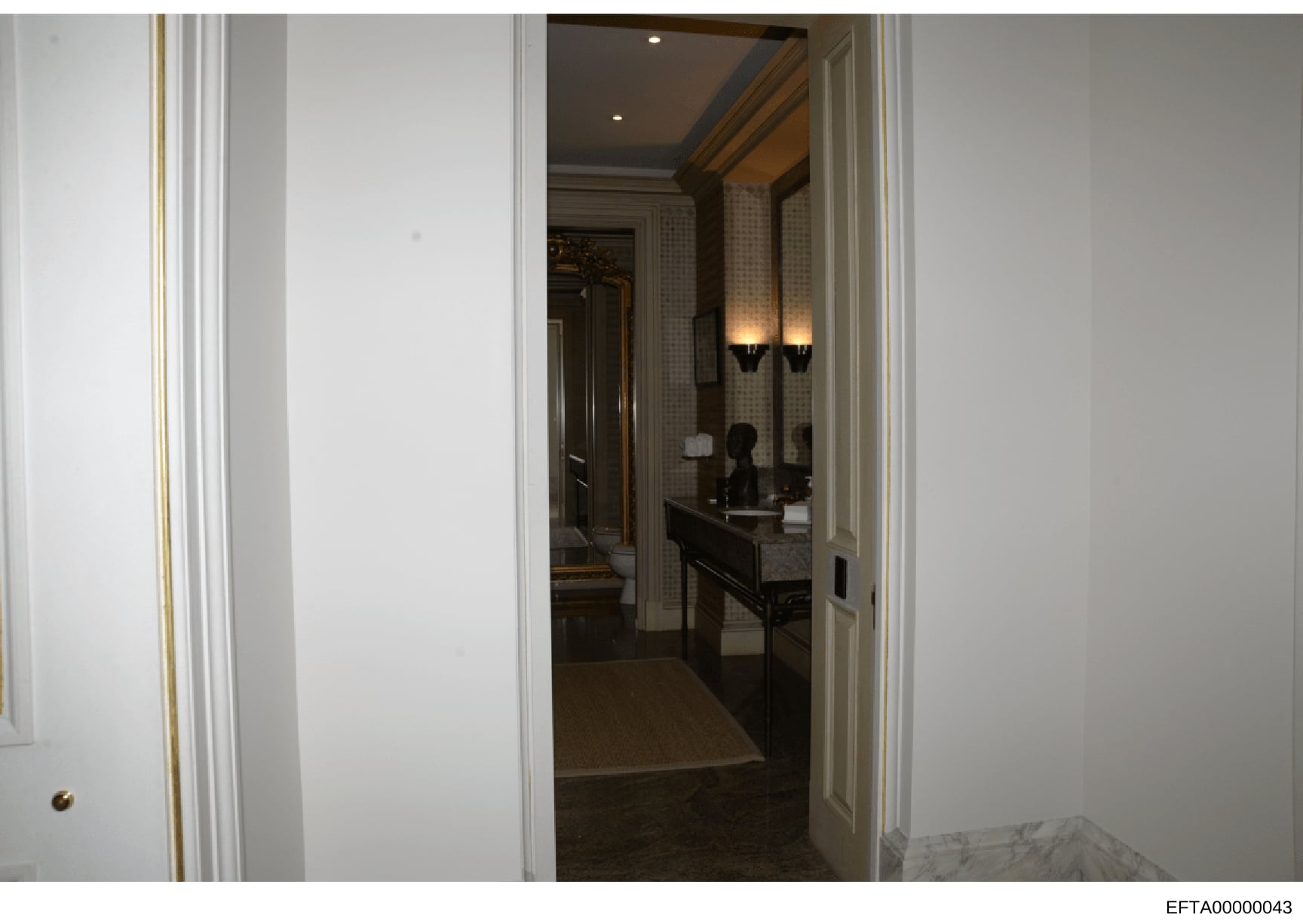 This photograph documents the interior of an upscale bathroom within a residence, showing a double vanity setup with sophisticated finishes, recessed lighting, and decorative wall sconces. The image captures the doorway entrance from an adjoining hal