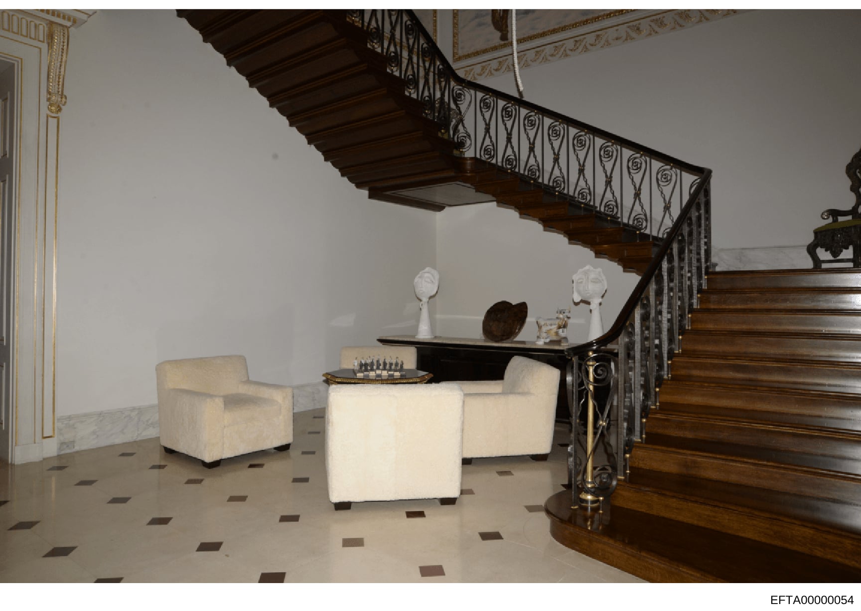 This photograph documents the interior of a luxurious residence, showing a multi-story foyer with an ornate wrought-iron staircase, cream-colored upholstered seating furniture, decorative sculptures, and marble flooring with inlaid pattern work. The 