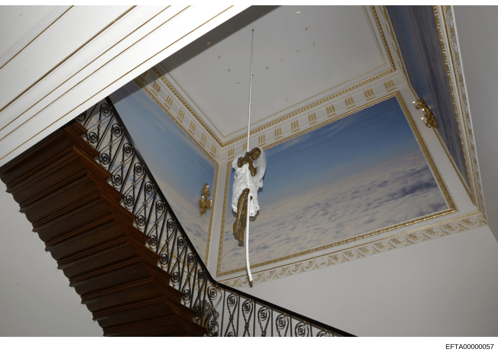 This is a photograph of an interior space featuring luxurious architectural details including an ornate ceiling with gold leaf trim, a framed cloud-sky mural, wrought iron staircase railing with decorative spirals, and chandelier elements. The image 