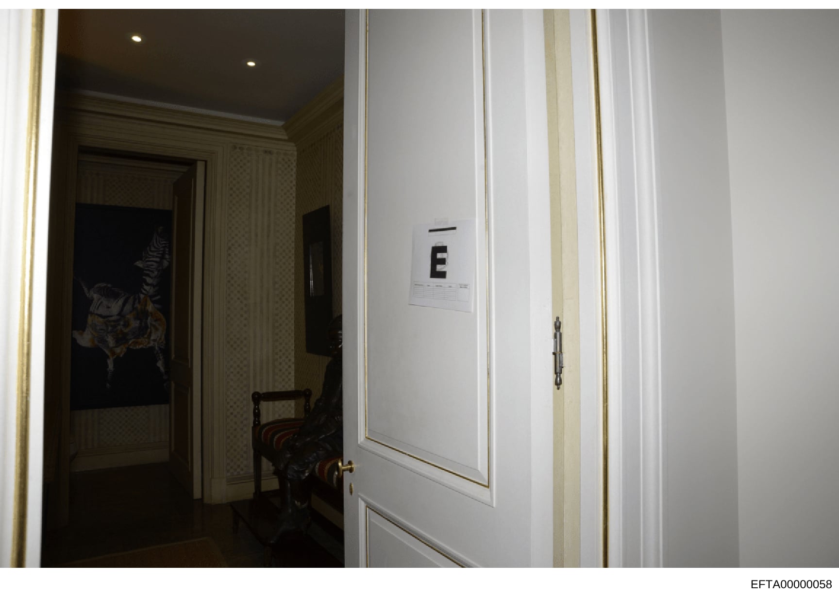 This is a photograph of an interior hallway in what appears to be an upscale residence, showing a white paneled door marked with the letter 'E' and a Bates stamp. The image documents the interior layout of a property, likely taken during an evidence 