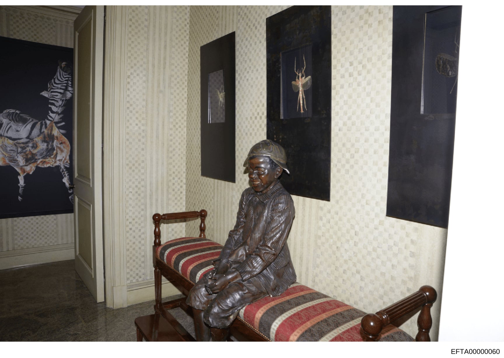This photograph depicts an interior room containing a bronze statue of a figure seated on a striped upholstered chair, surrounded by framed artwork featuring wildlife subjects including zebras and deer. The room features patterned wallpaper and appea