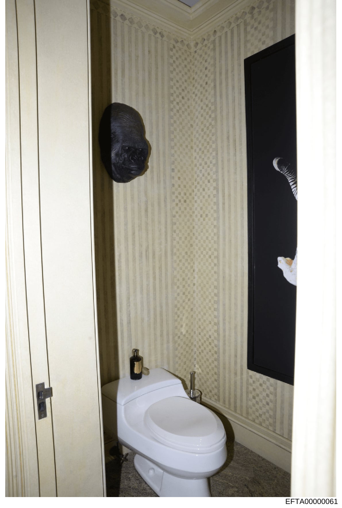 This is a photograph of a small bathroom interior, showing a white toilet, wallpapered walls with a subtle pattern, and notably featuring two pieces of dark art on the walls—a sculptural black mask or face on the left and what appears to be a black p