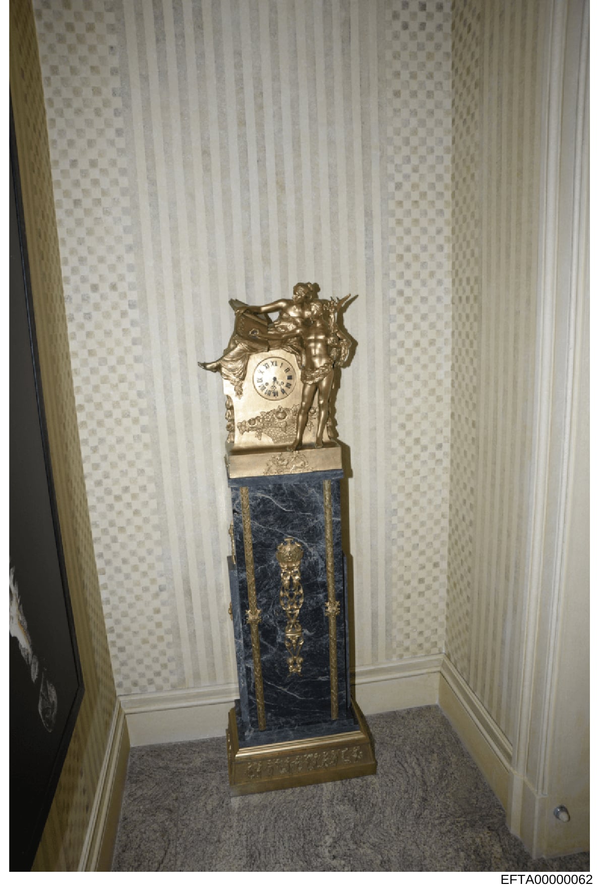 This photograph documents a decorative ornamental clock seized as evidence in the Epstein investigation. The clock features a bronze cherub and decorative elements mounted atop a dark blue marble pedestal with gilt bronze accents, photographed in wha