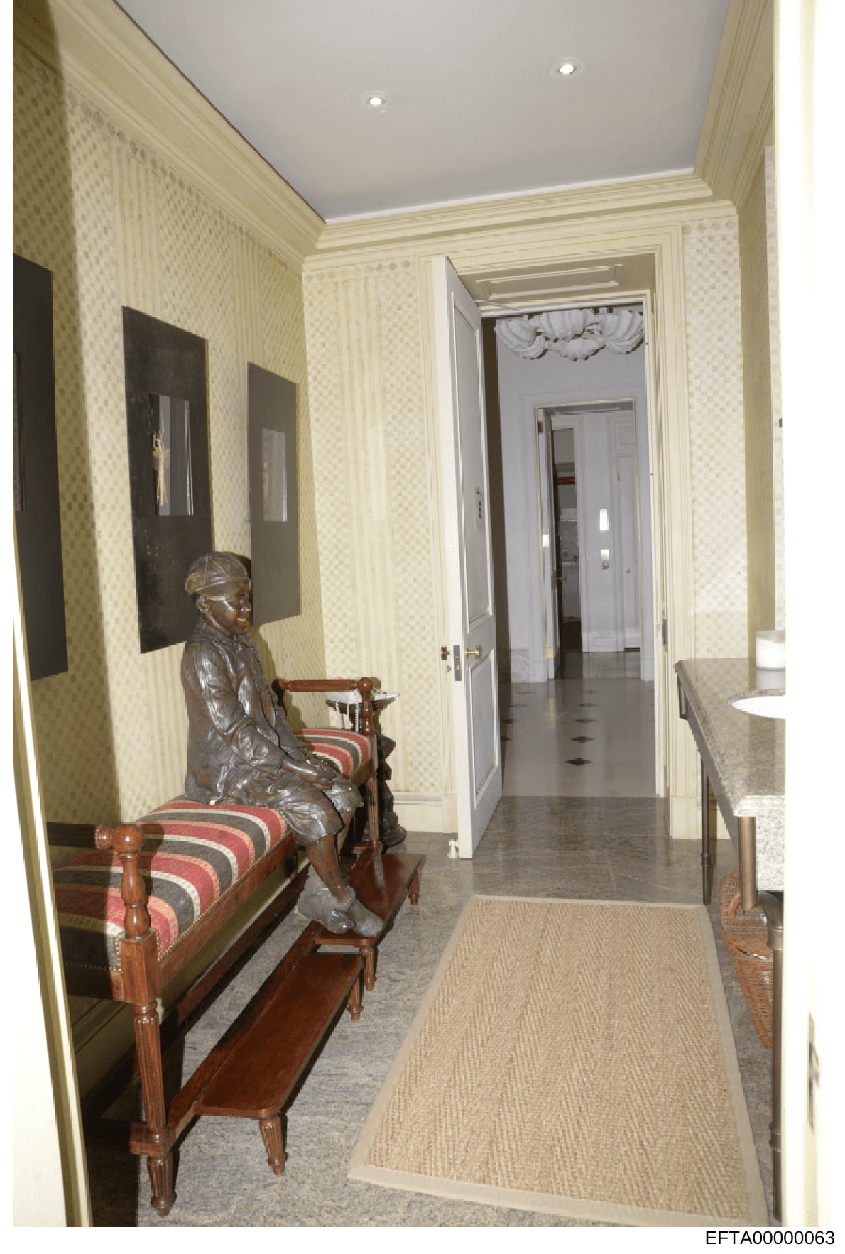 This photograph documents an elegantly decorated hallway within a residential property, featuring cream-colored damask wallpaper, recessed ceiling lighting, framed artwork, period furniture including a wooden bench with striped upholstery, and a bron