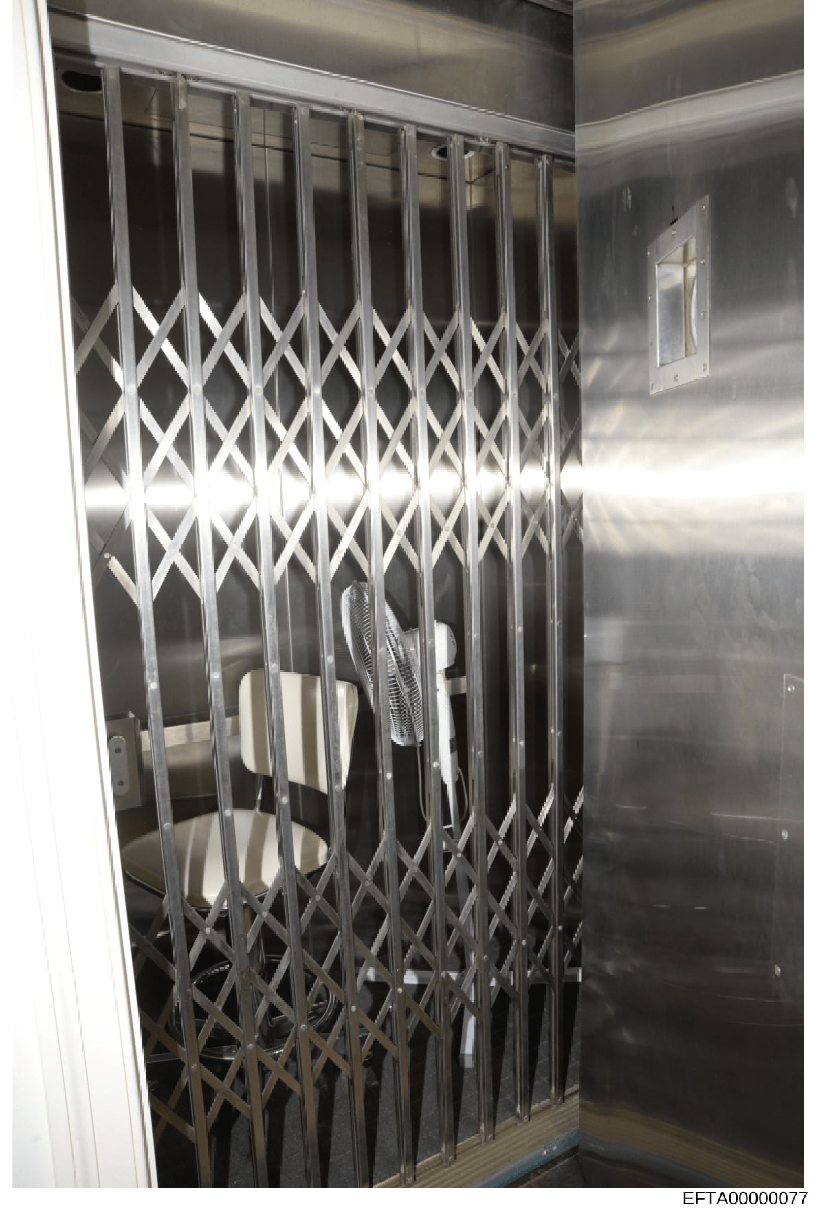 This is a photograph of an interior space featuring a closed accordion-style security gate with a diamond crosshatch pattern in white/light metal. The room has metallic or steel-colored walls and appears to be a secured or restricted area. The image