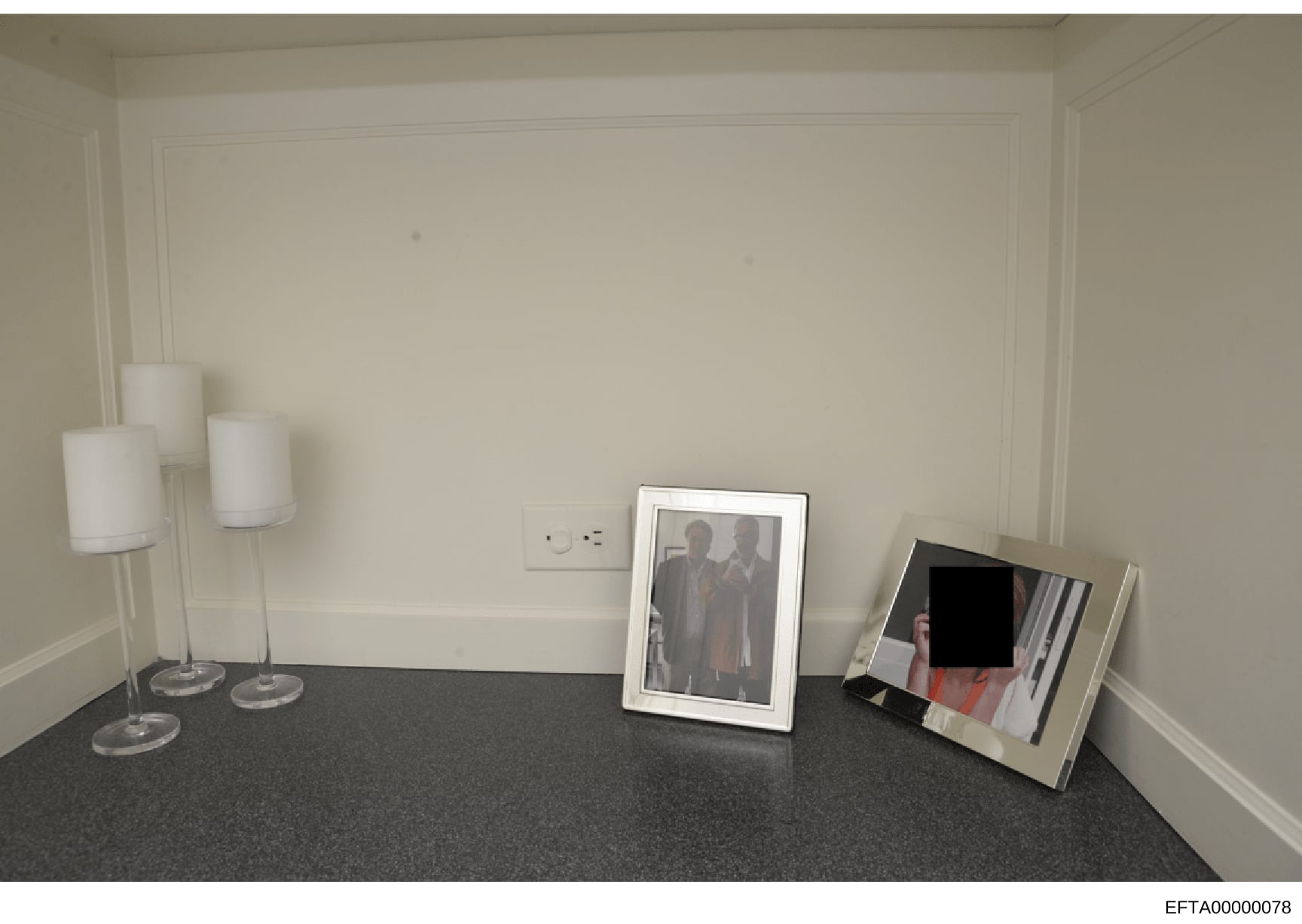 This is an evidence photograph showing the interior of a room with minimal furnishings. The image depicts a dark carpeted floor, cream-colored walls with wall sconce lighting, and three framed photographs placed on the floor against the wall. Two pho