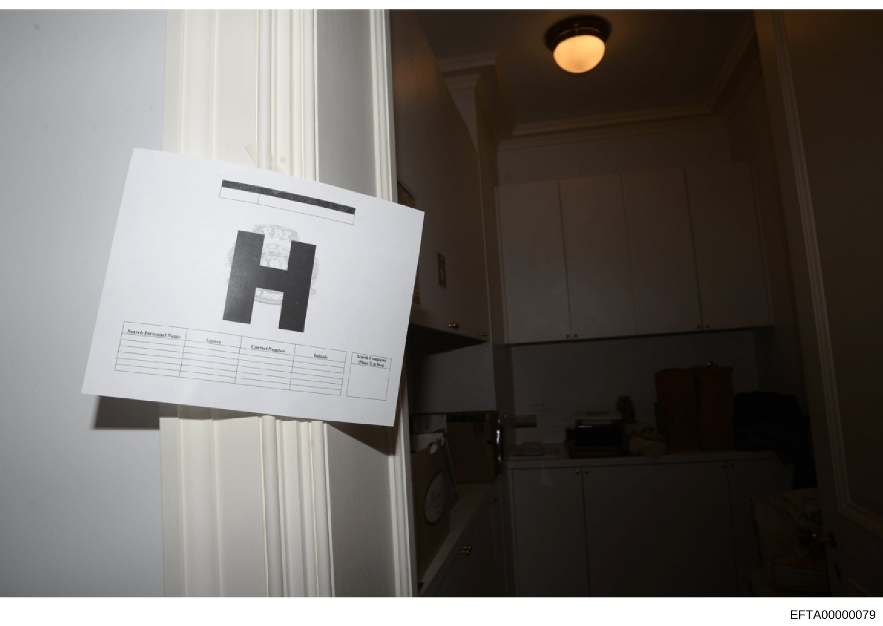This is an evidence photograph taken during a search warrant execution, showing a doorway or entrance with an evidence scale marker (marked with 'H') affixed to document paper. The photo documents a specific location within a property during the inve