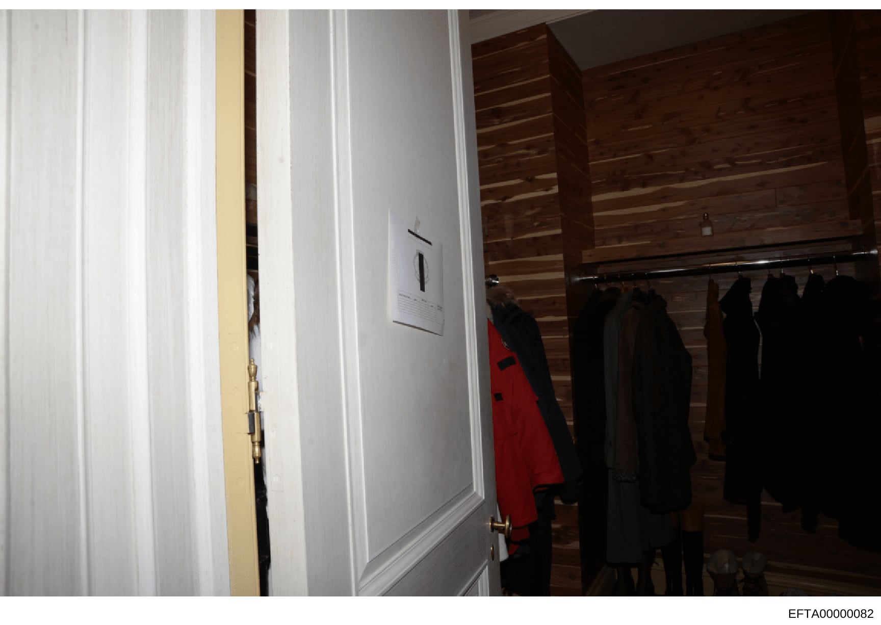 This is an evidence photograph showing an interior closet or dressing room space containing multiple dark-colored garments hanging on a rod, with a red jacket visible among them. The space features white paneling, wooden cabinetry, and wall fixtures.