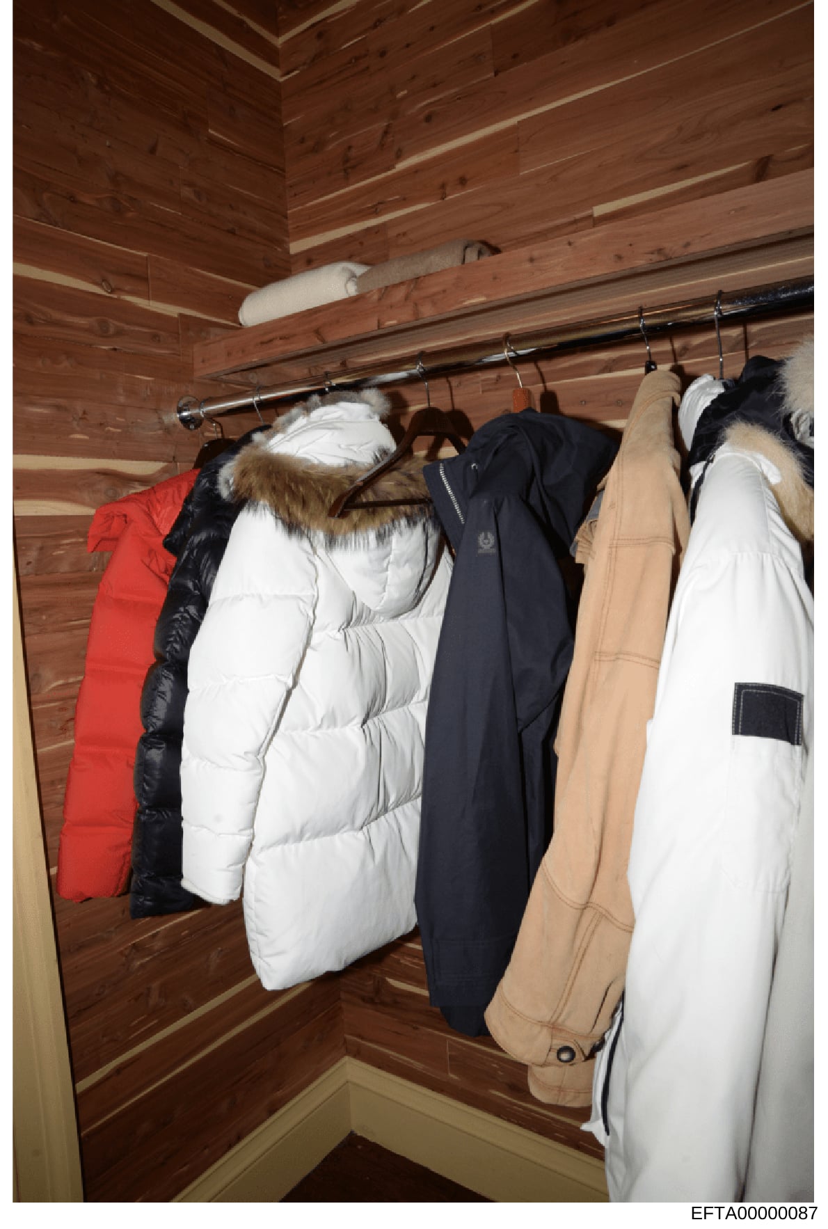 This is a photograph of a closet interior with wood-paneled walls and ceiling. The closet contains multiple winter coats and jackets hanging on a chrome rod, including white puffy down jackets, a red jacket, a black jacket, and tan/beige colored oute