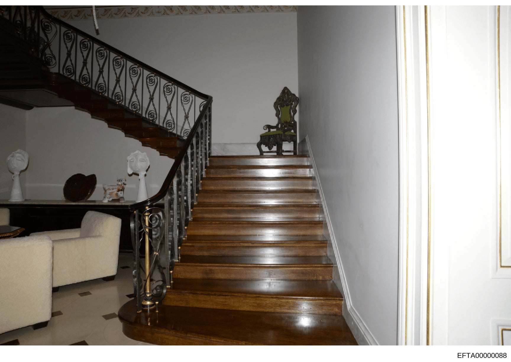 This photograph shows the interior of a luxurious residence, depicting a grand wooden staircase with ornate wrought-iron railings, polished hardwood steps, and elegant architectural details. The image includes period furniture and decorative objects 