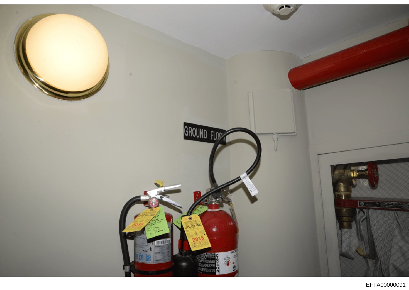 This photograph documents fire safety equipment located on the ground floor of one of Epstein's properties. The image shows two fire extinguishers mounted on a wall, labeled with inspection tags dated 2018, suggesting routine maintenance records. The
