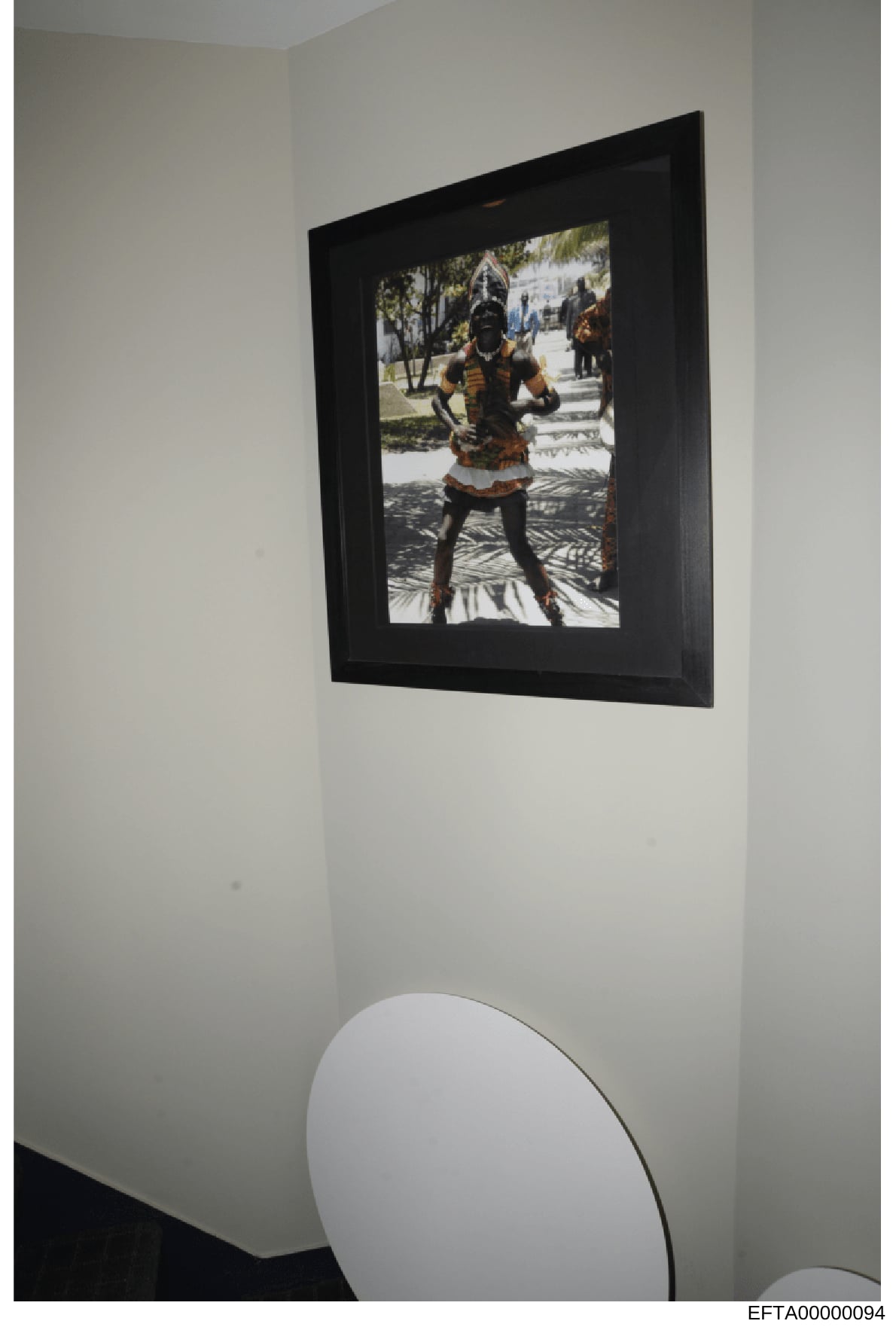 This is a photograph of a framed artwork hanging on a white interior wall. The framed image depicts a person in traditional African dress with beaded accessories and body paint, photographed outdoors with trees and white buildings visible in the back