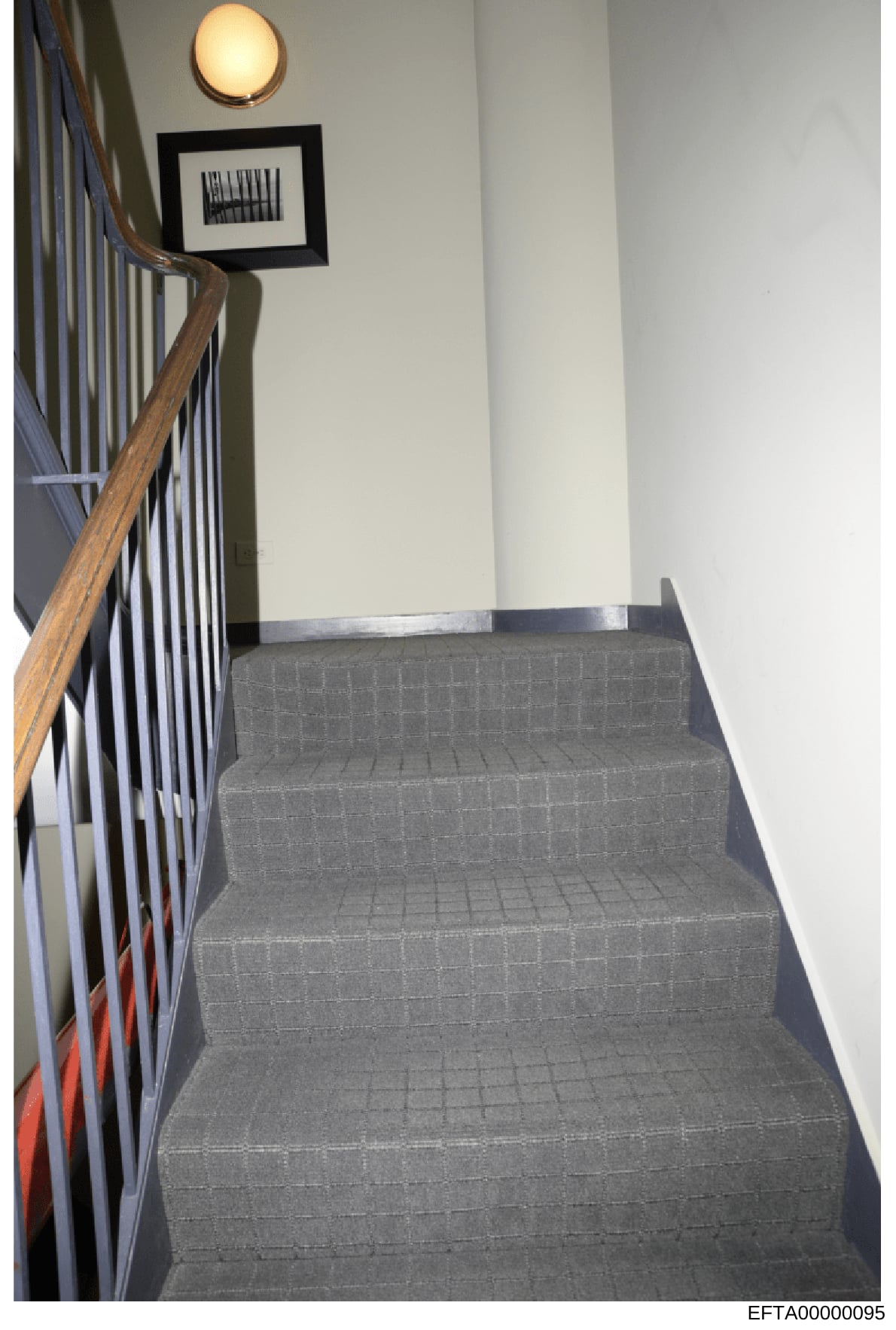 This photograph shows an interior staircase from one of Jeffrey Epstein's properties, featuring gray carpeted stairs, wooden handrails, a framed artwork on the wall, and decorative lighting. The image provides documentation of the interior design and