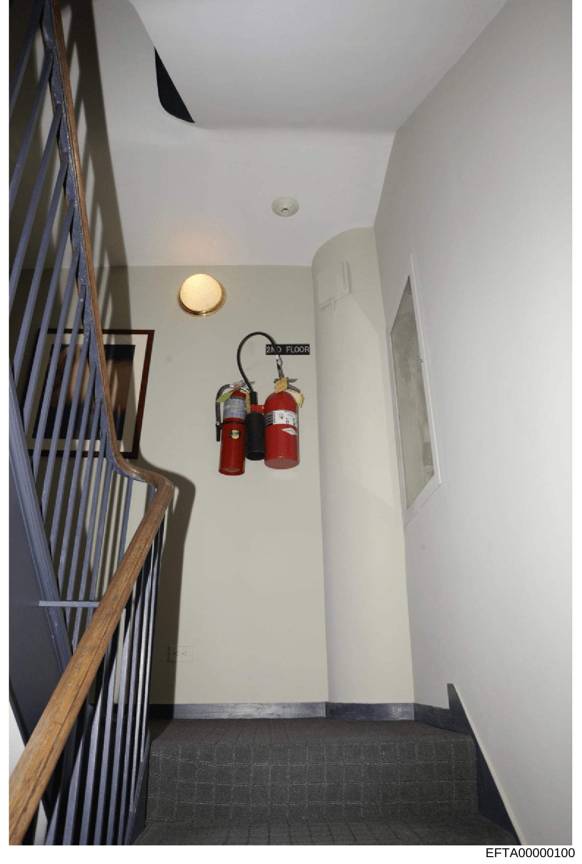 This photograph depicts an interior stairwell landing on the second floor of a building, showing safety equipment and architectural features. The image documents the stairwell infrastructure including handrails, lighting fixtures, and fire extinguish