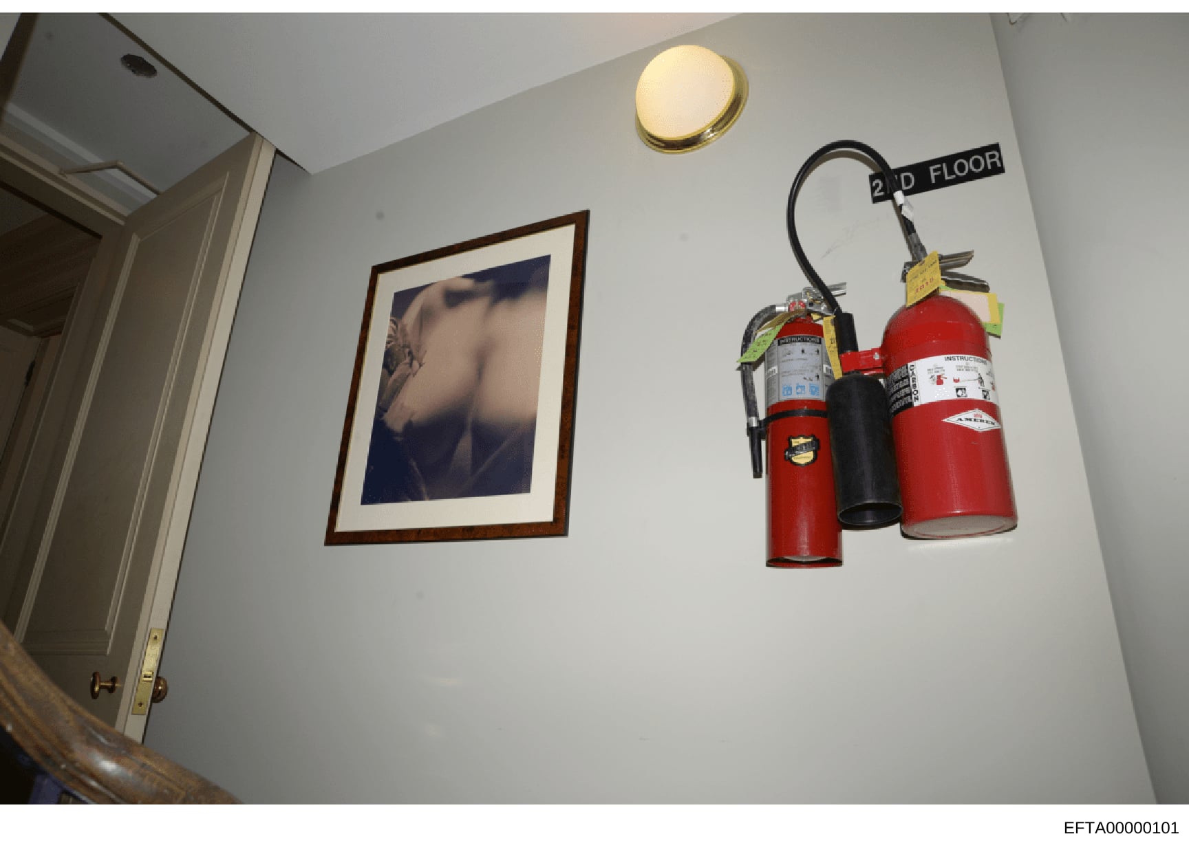 Evidence photo showing the interior of a second-floor room, featuring wall-mounted fire extinguishers, a framed artistic photograph, ceiling light fixture, and doorway. This appears to be a documentary photo from the investigation premises, likely ta