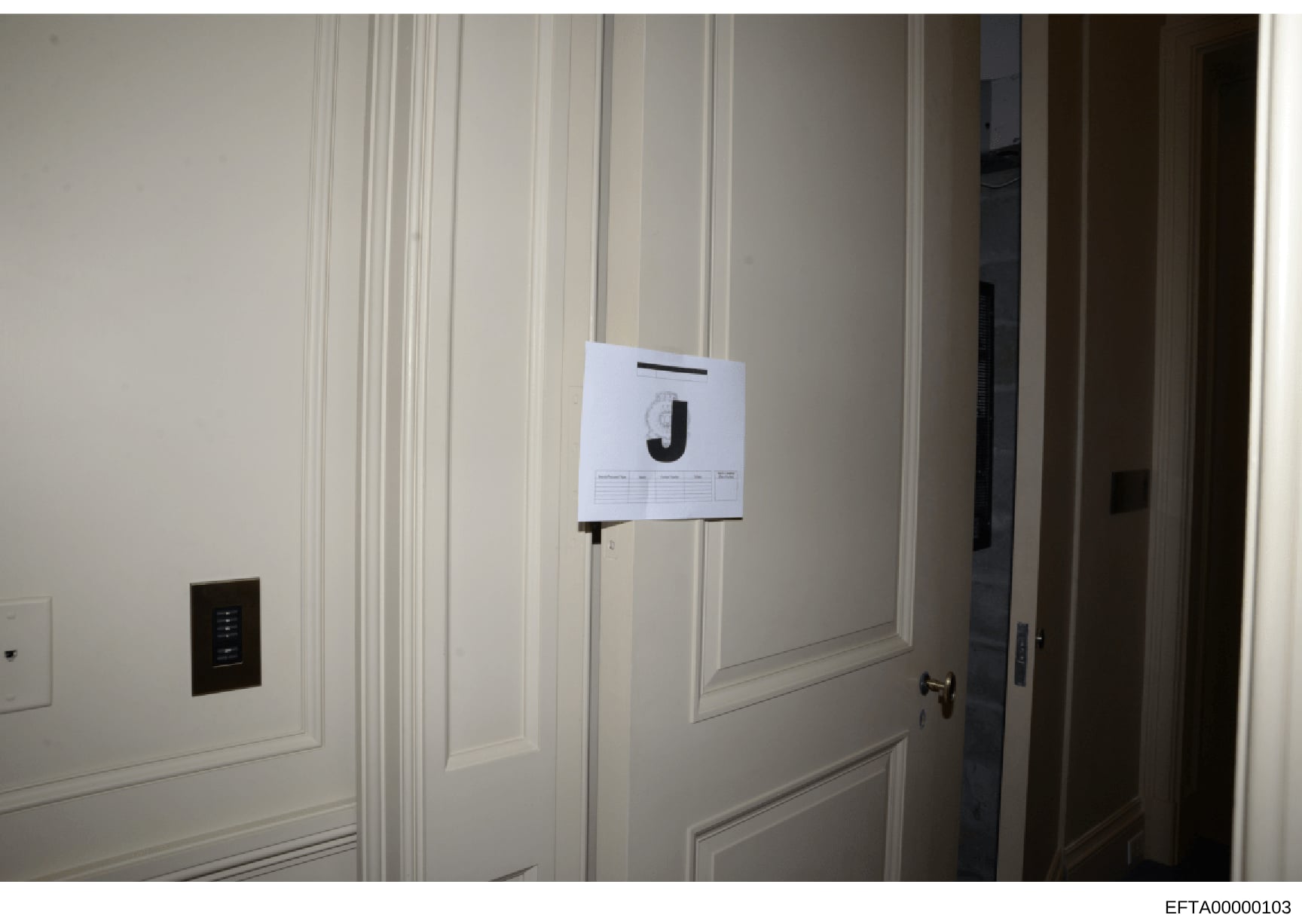This is an evidence photograph from the Epstein investigation showing an interior room with cream-colored paneled walls, a dark electrical panel on the left, and a large white evidence marker labeled 'J' mounted on the wall. The photo appears to be p