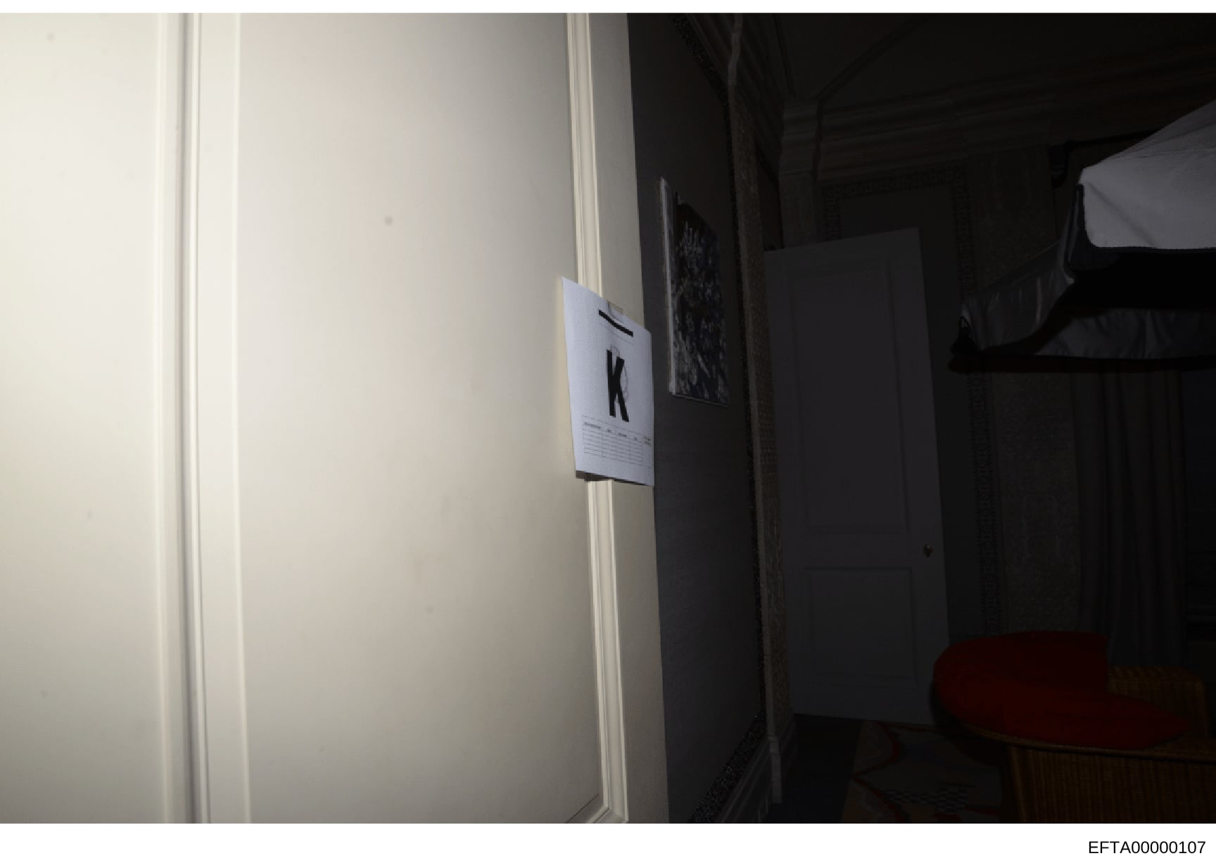 This is an evidence photograph showing an interior room with a white label marker bearing the letter 'K' affixed to a door frame or wall. The photo appears to be taken during a property search or investigation, with the marker used to identify and ca