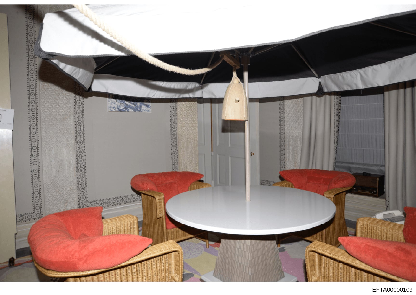 This photograph depicts an interior space featuring a modern design setup with a round white table, multiple wicker chairs with red cushions arranged in a circular configuration, and an elaborate canopy structure with black and white striped fabric o