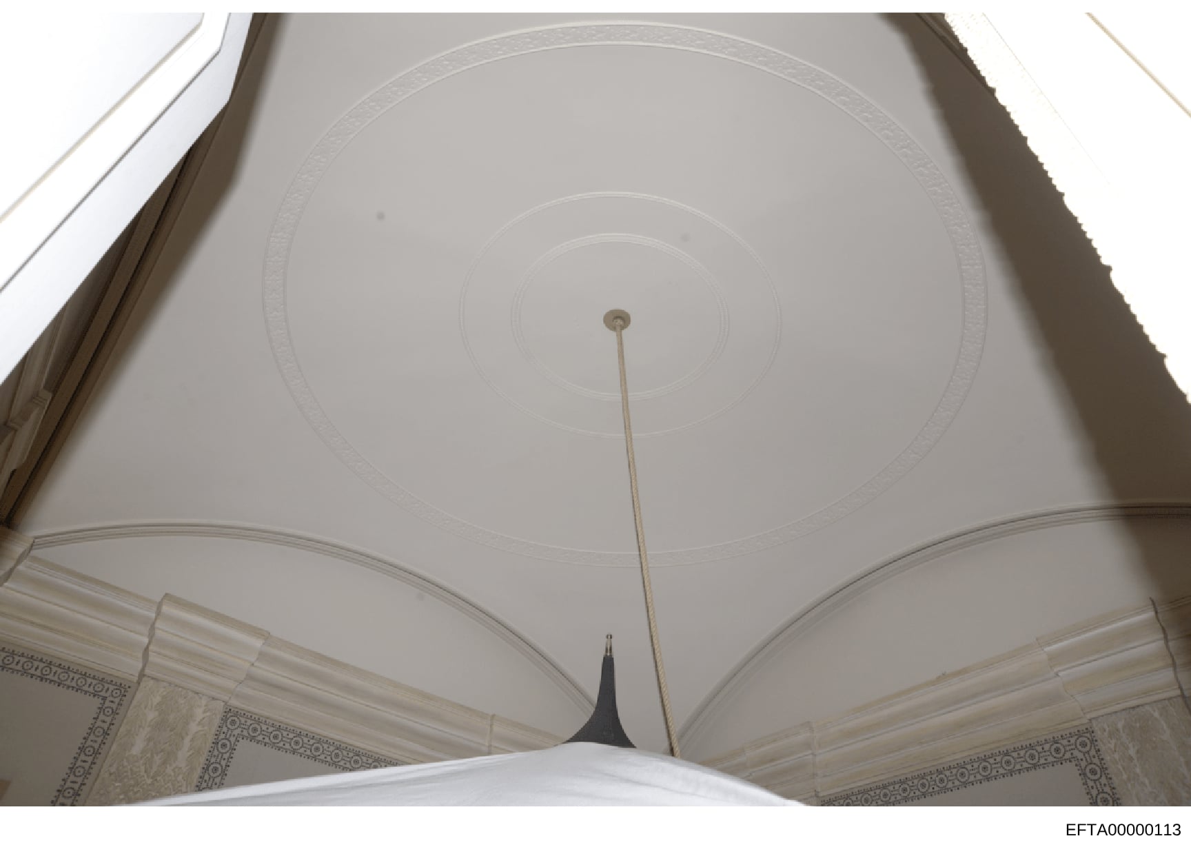 This is a photograph of an interior ceiling featuring classical architectural elements including a domed or barrel-vaulted ceiling with concentric circular details, decorative molding, and ornamental borders on the walls. The image appears to be take