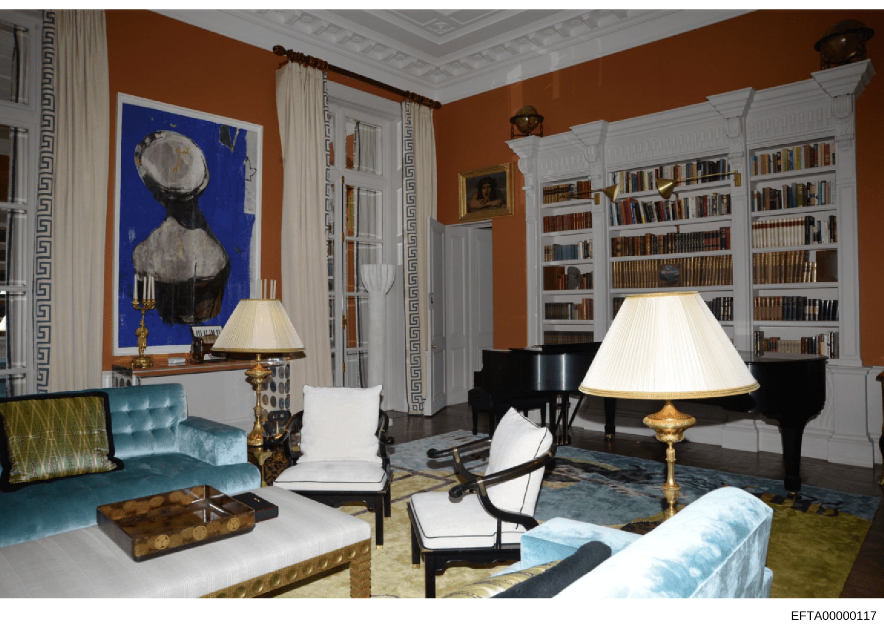 This is a photograph of an elegantly decorated formal living room featuring sophisticated furnishings, extensive built-in bookshelves, a grand piano, and fine art. The room displays classic interior design with warm orange walls, white architectural