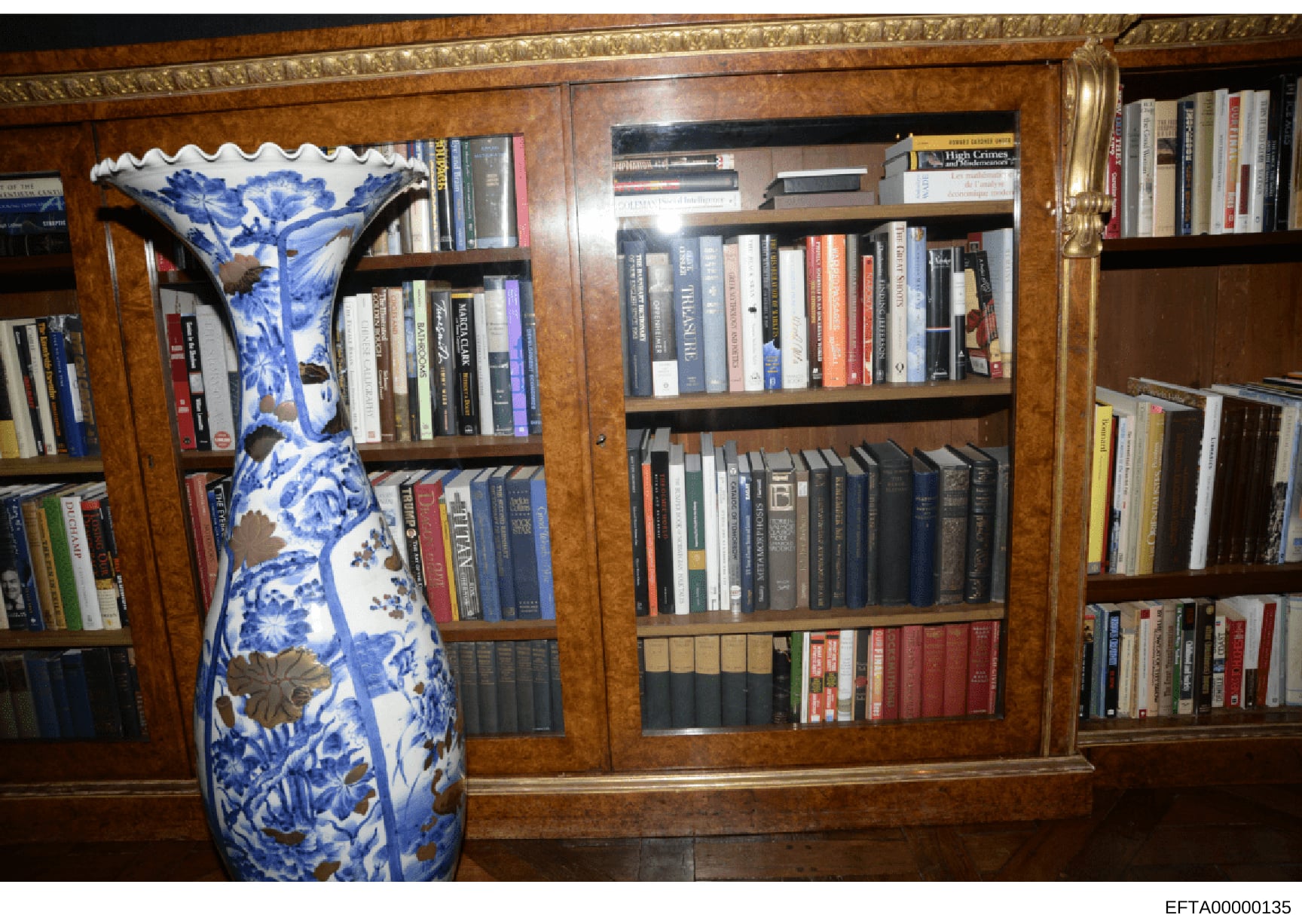 This photograph shows the interior of a residence, depicting a large wooden bookcase filled with books and a prominent blue and white porcelain decorative vase. The image appears to be evidence documentation from the Epstein investigation, likely tak