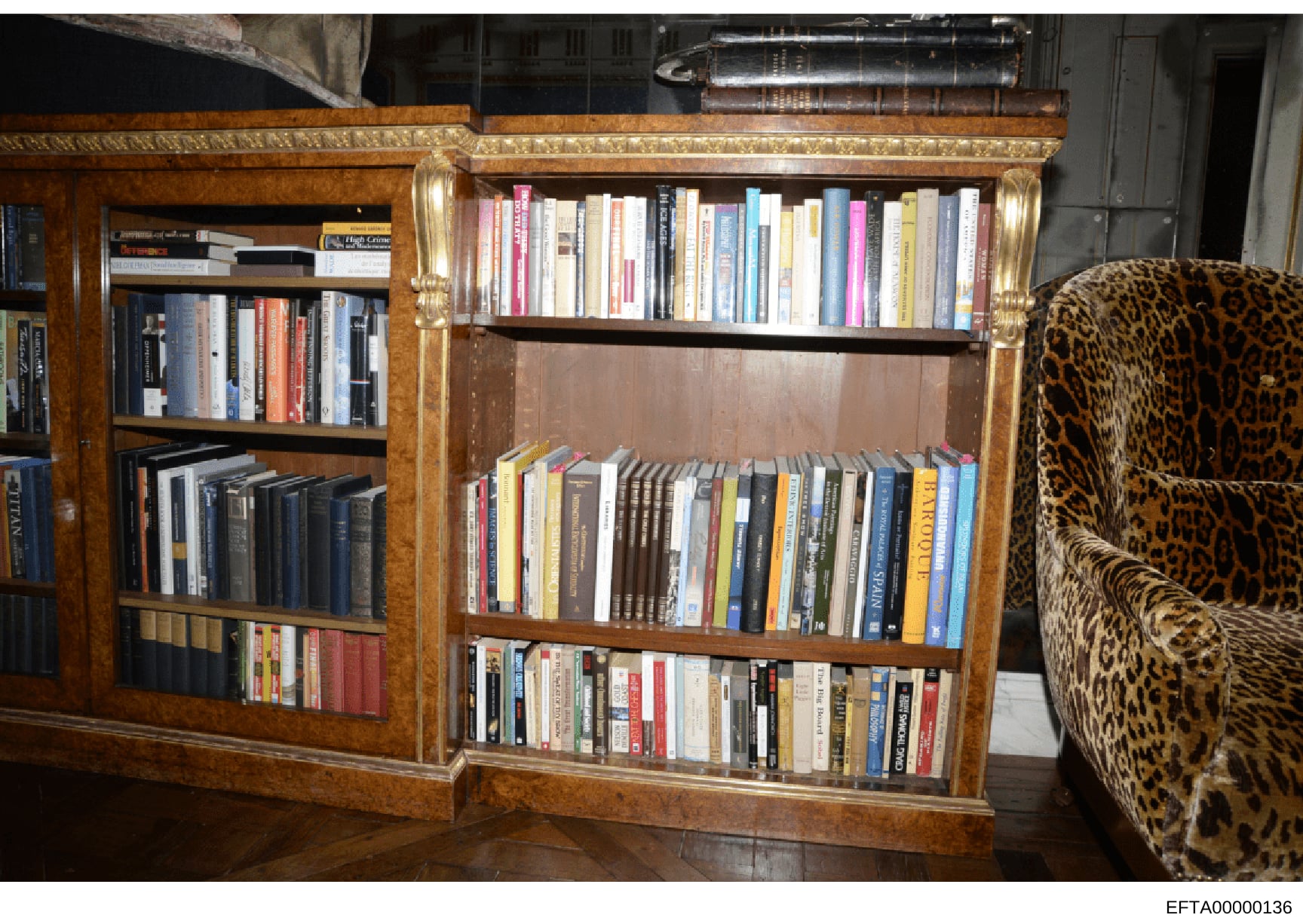 This is a photograph of an interior room at one of Jeffrey Epstein's properties, showing an ornate wooden bookcase unit with glass doors containing hundreds of books, alongside a leopard-print upholstered chair. The image documents the furnishings an