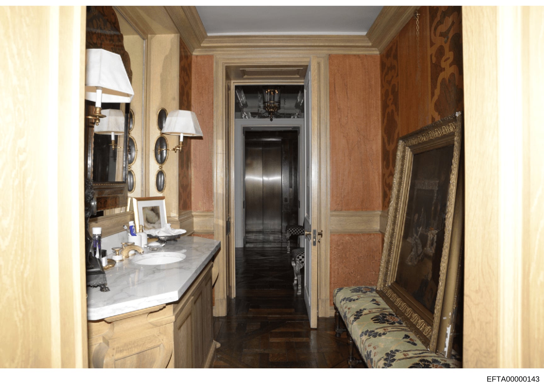 This is a photograph of an elegantly appointed bathroom and adjoining hallway from what appears to be a high-end residential property. The image shows custom marble countertops, brass fixtures, damask wallpaper, ornate mirrors, and decorative furnish