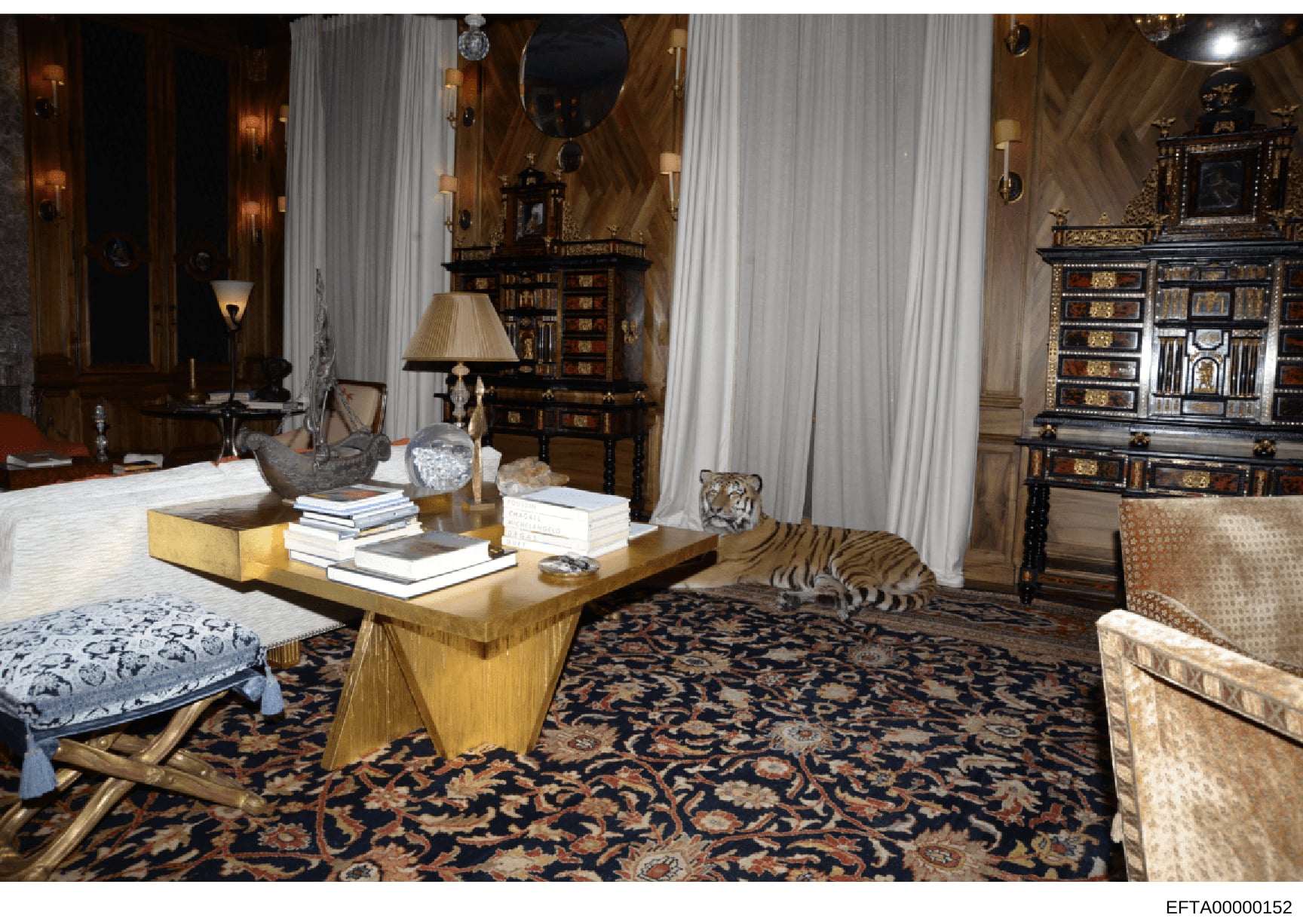 This photograph depicts an elegantly furnished formal living room or parlor featuring high-end furniture, artwork, and decorative elements. The room includes antique cabinets, a gold-leaf coffee table with books, ornate mirrors, wall sconces, and dec