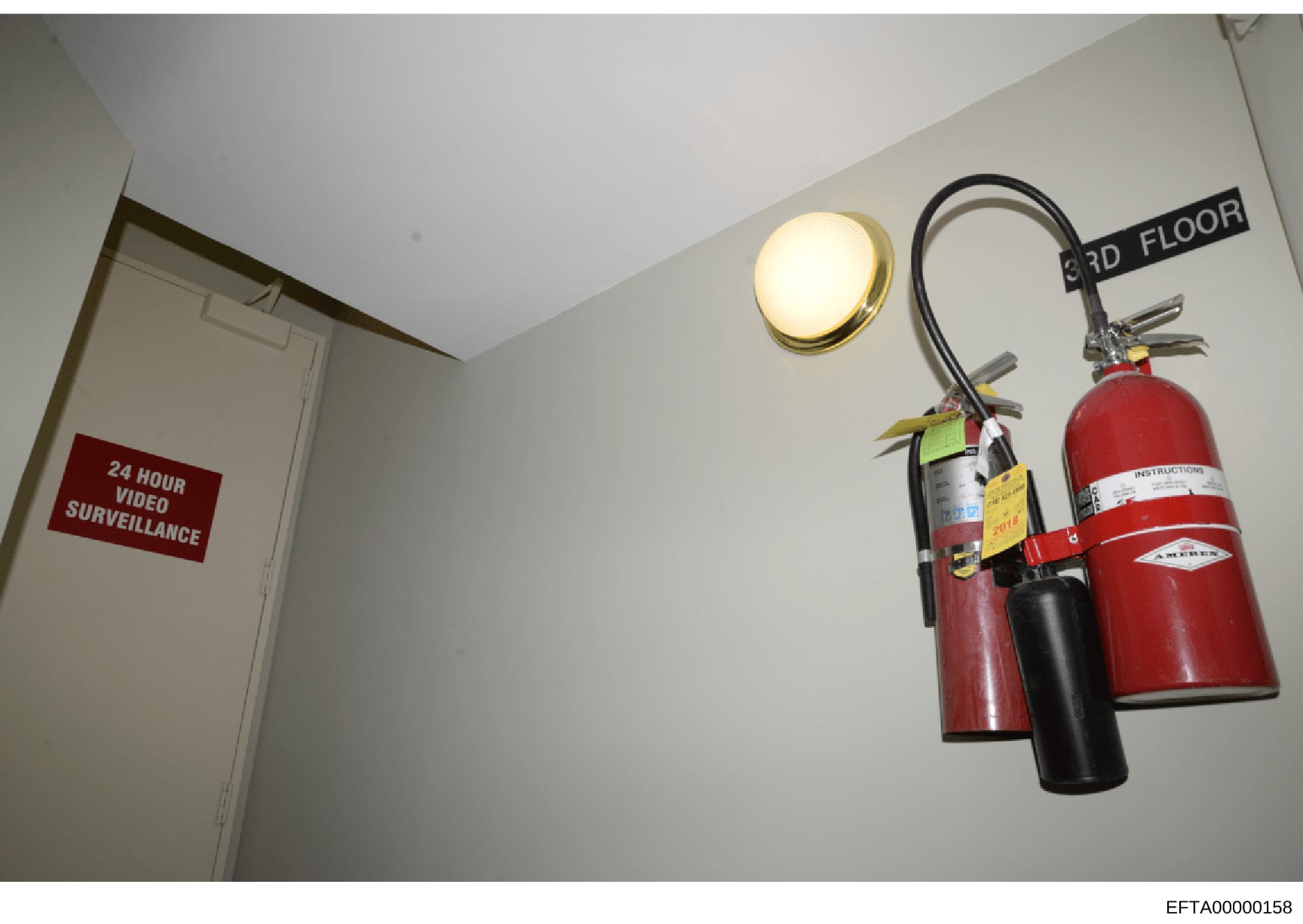 This is an evidence photograph from an Epstein property showing a third-floor hallway with a prominent '24 HOUR VIDEO SURVEILLANCE' sign on a door, a ceiling-mounted light fixture, and fire safety equipment including multiple fire extinguishers mount