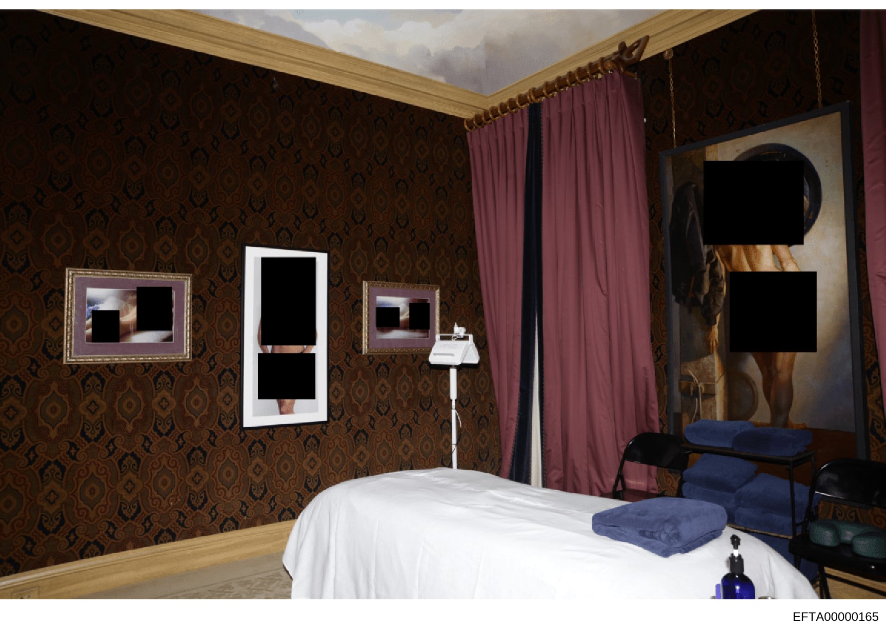 This photograph depicts an interior bedroom space featuring ornate décor, including damask-patterned wallpaper in brown and gold tones, burgundy curtains, framed artwork with redacted content, a white bed with blue cushions, and various furnishings. 