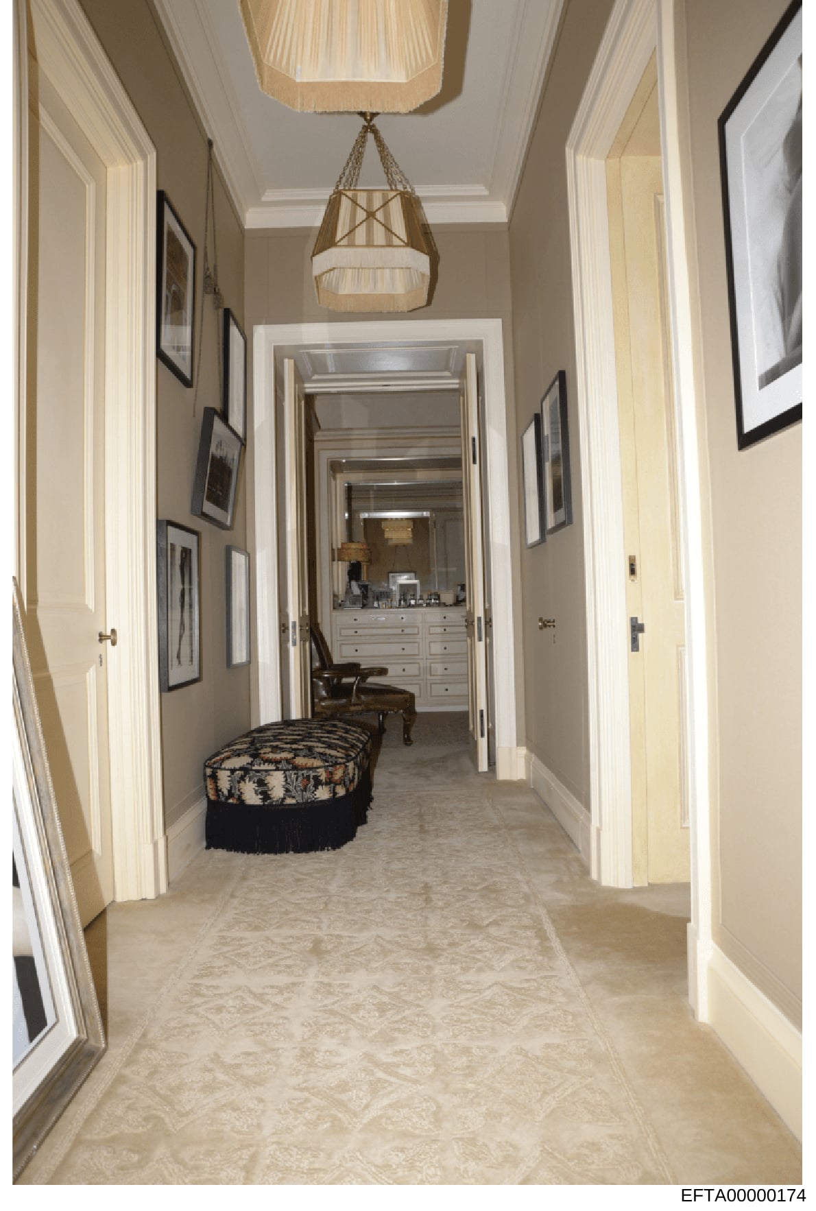 This is a photograph of an elegant residential hallway, featuring cream and beige tones, framed artwork on the walls, a decorative pendant light fixture, and a view into a dressing room with white built-in cabinetry and vanity. The image documents in