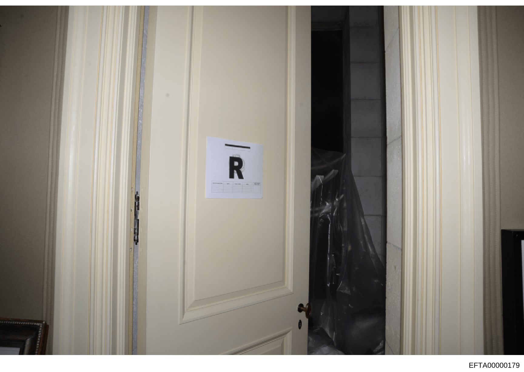 This is a photograph of an interior room showing a cream-colored wall with vertical molding and a doorway. A white evidence marker labeled 'R' is positioned on the wall. A black plastic evidence bag is visible in the doorway behind the marker. The ph