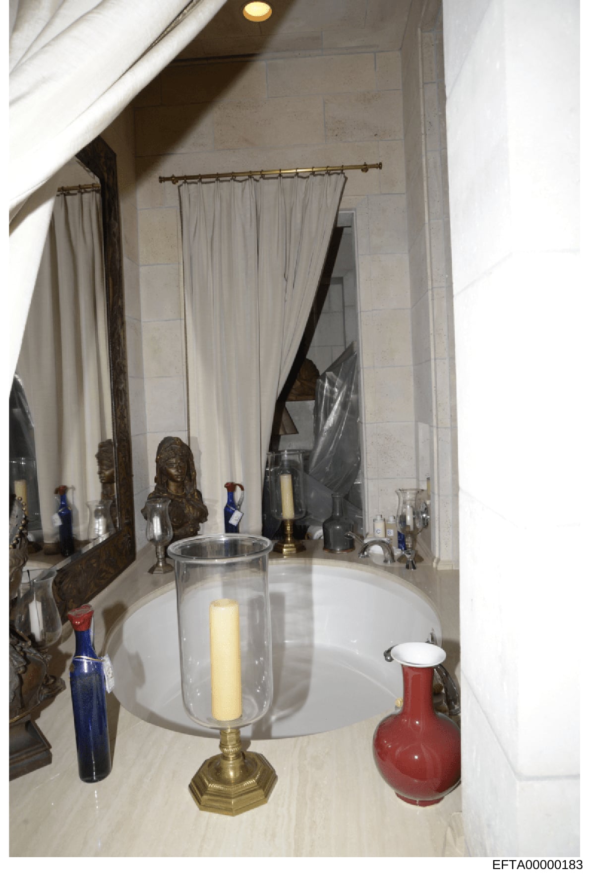 This is a photograph of an upscale bathroom interior, showing a marble countertop with decorative items including a brass candle holder, bronze bust sculpture, colored glass bottles, and a red ceramic vessel. The image documents the interior design a