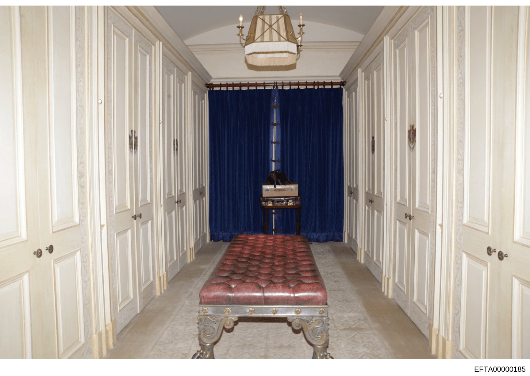 This is a photograph of a long, formal hallway in what appears to be a luxury residence, featuring white paneled closet doors on both sides, a red upholstered bench in the center, deep blue curtains at the far end, and decorative architectural elemen