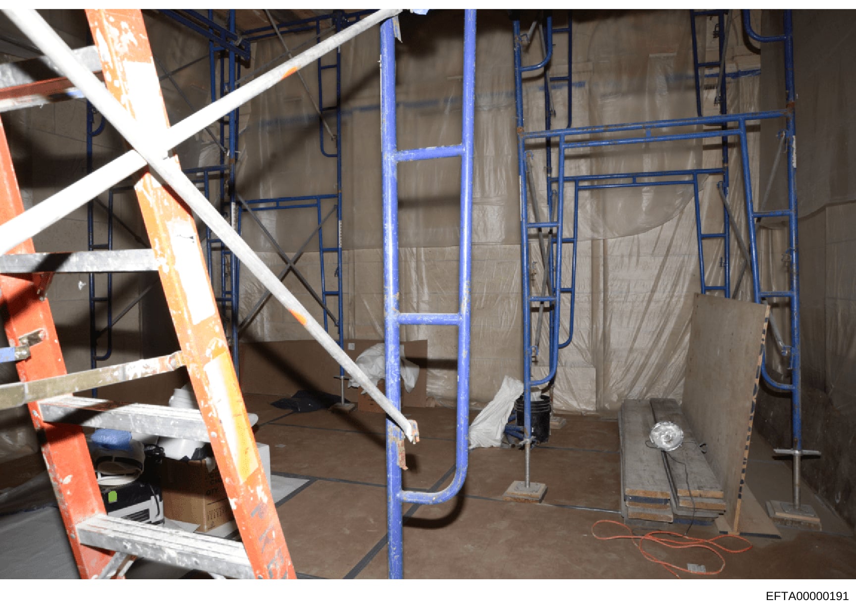 This photograph shows an interior space under active construction or renovation, featuring blue metal scaffolding, protective plastic sheeting, an orange ladder, and construction equipment. The image appears to be documentation of a property renovati