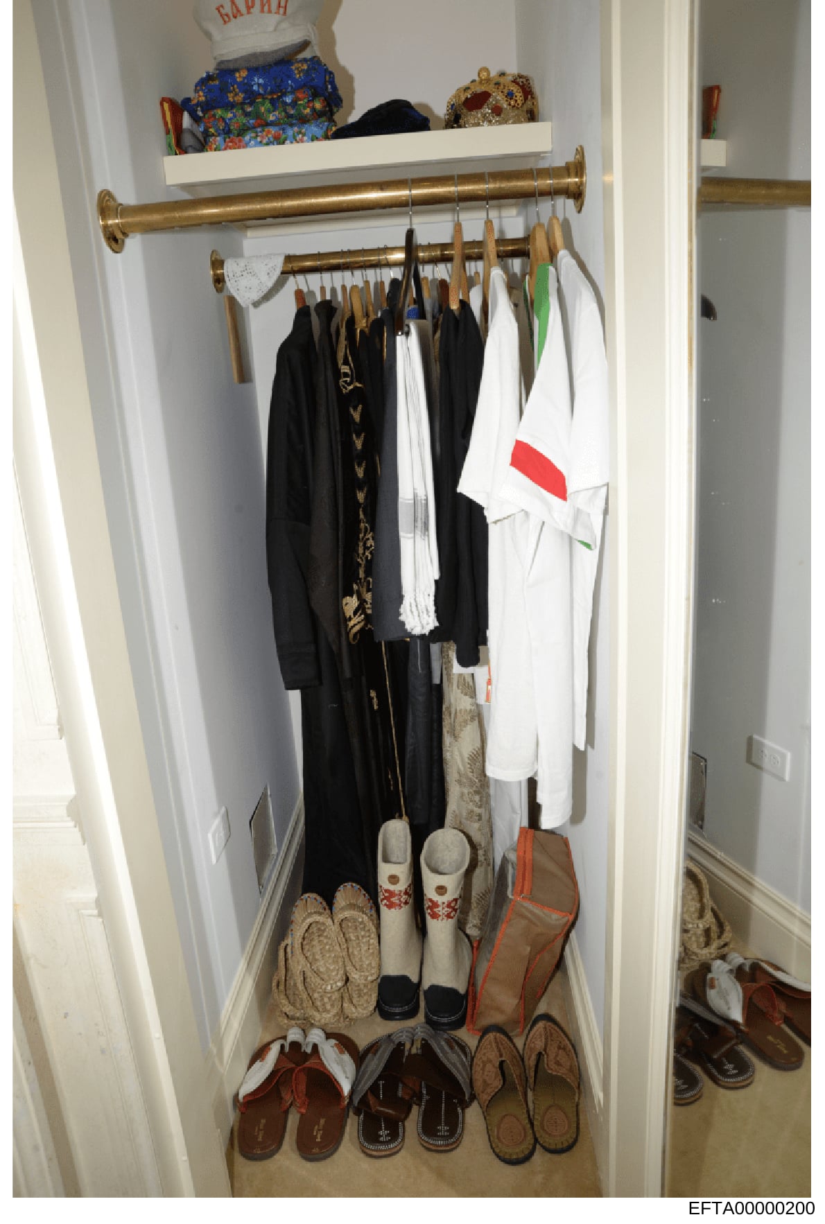 This is a photograph of a residential closet interior containing clothing, shoes, and personal items. The closet features brass rods for hanging garments including black and white clothing, and the floor displays numerous pairs of slippers and shoes