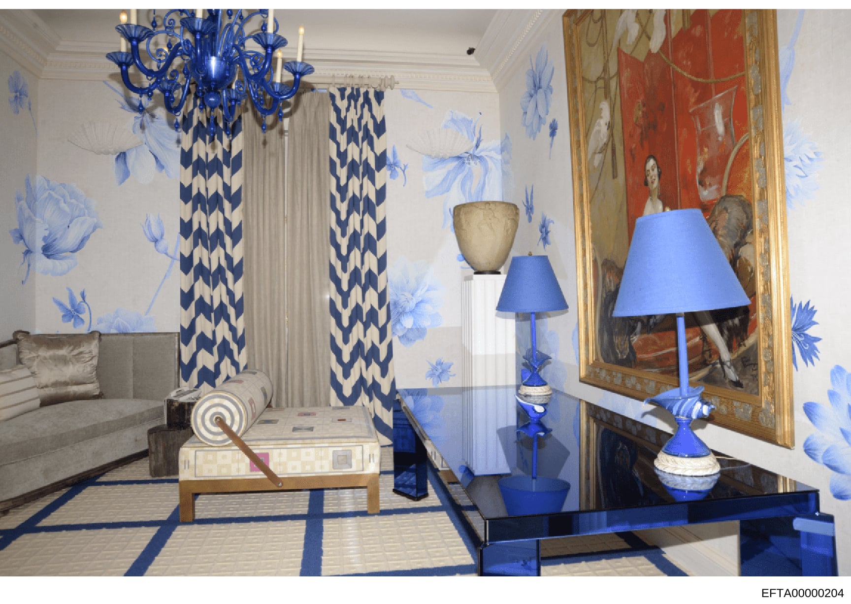 This is a photograph of an elegantly decorated interior space featuring a sophisticated blue and white color scheme. The room displays luxury furnishings including a blue Murano-style chandelier, patterned curtains, fine art, decorative lamps, a glas