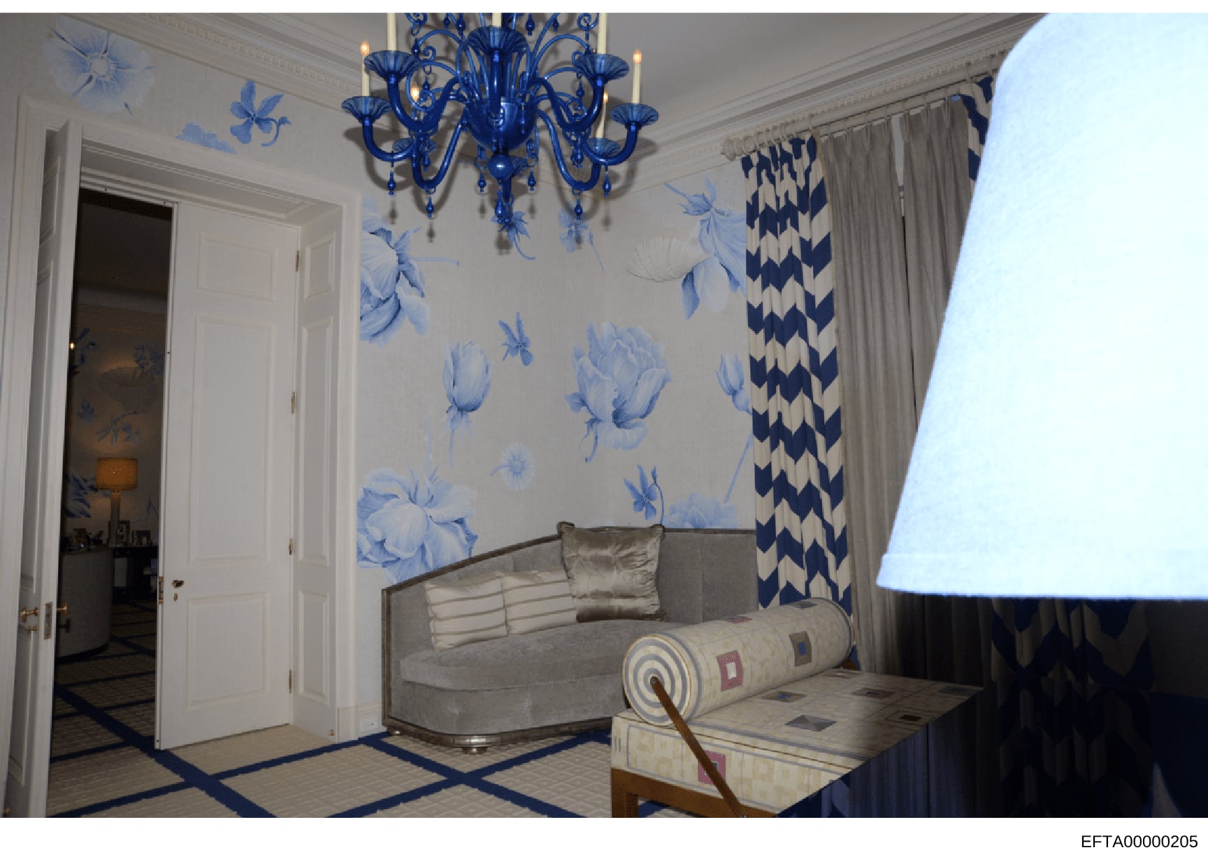 This photograph shows an elegantly decorated interior room featuring blue and white décor, including a distinctive blue Murano glass chandelier, floral wallpaper, decorative curtains, and upholstered furniture. The room appears to be part of a high-e