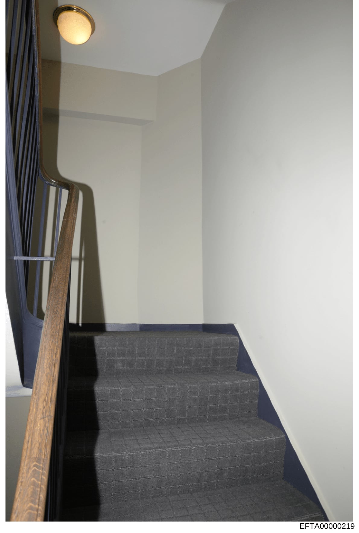 This is an evidence photograph showing an interior stairwell with modern finishes, including dark carpeted steps, light-colored walls, wooden handrail, and recessed lighting. The photo appears to be part of property documentation in the investigation